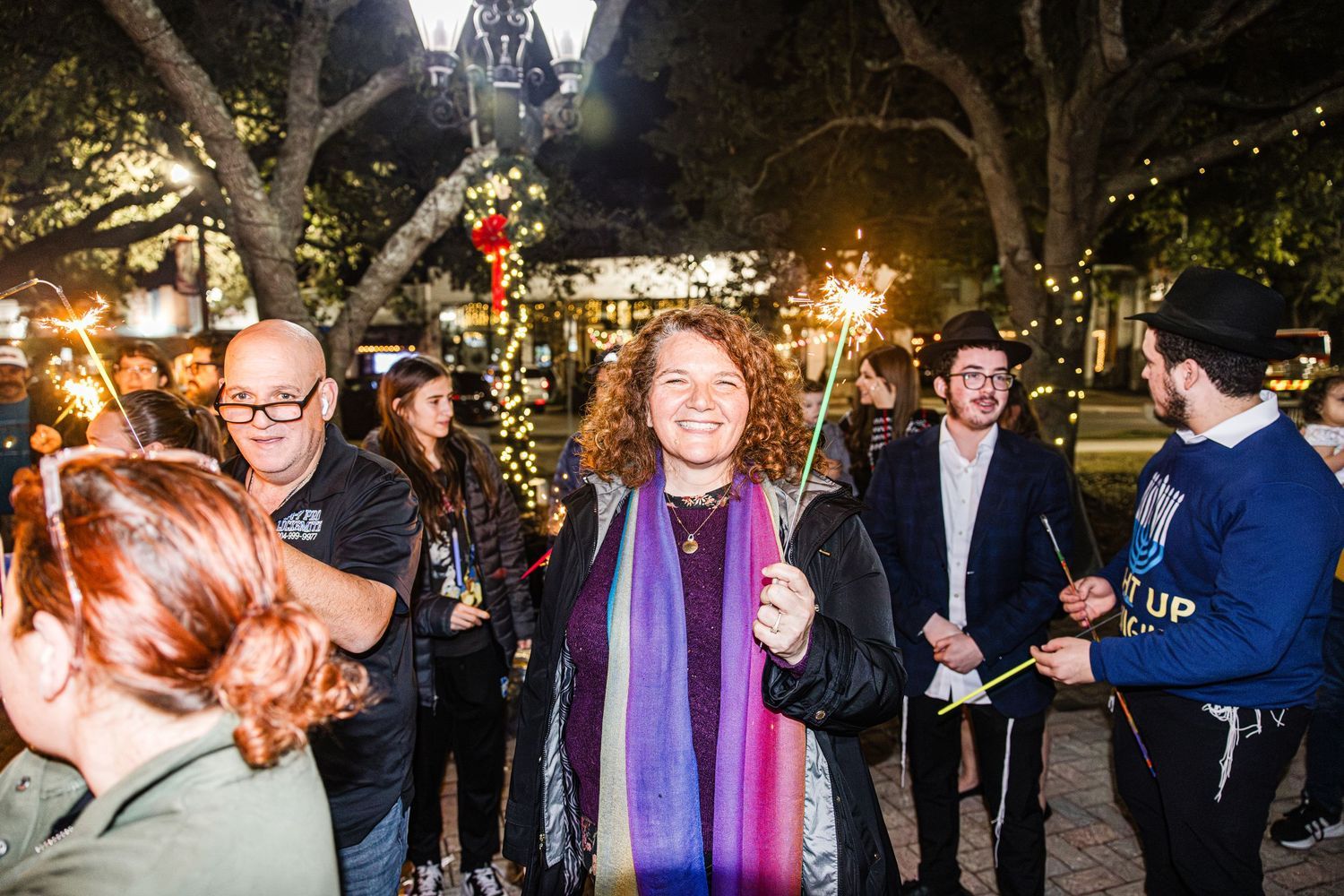 Woman in purple scarf smiles at outdoor Hanukkah celebration with sparklers at night.