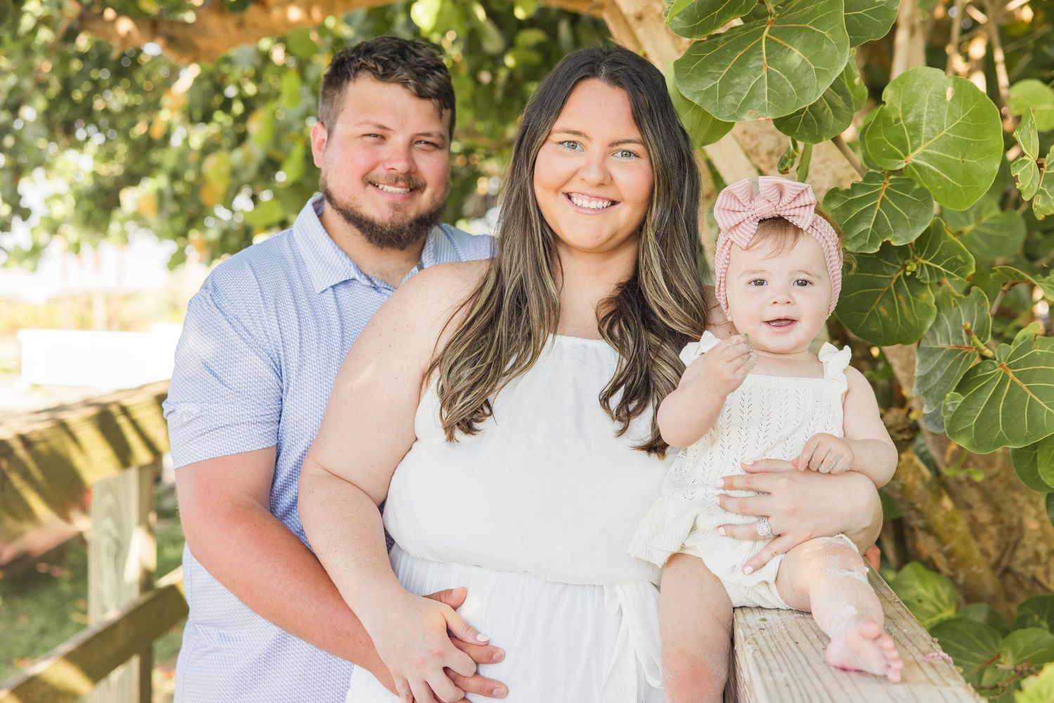 Young couple with baby girl in pink bow headband posing under tropical foliage outdoors.