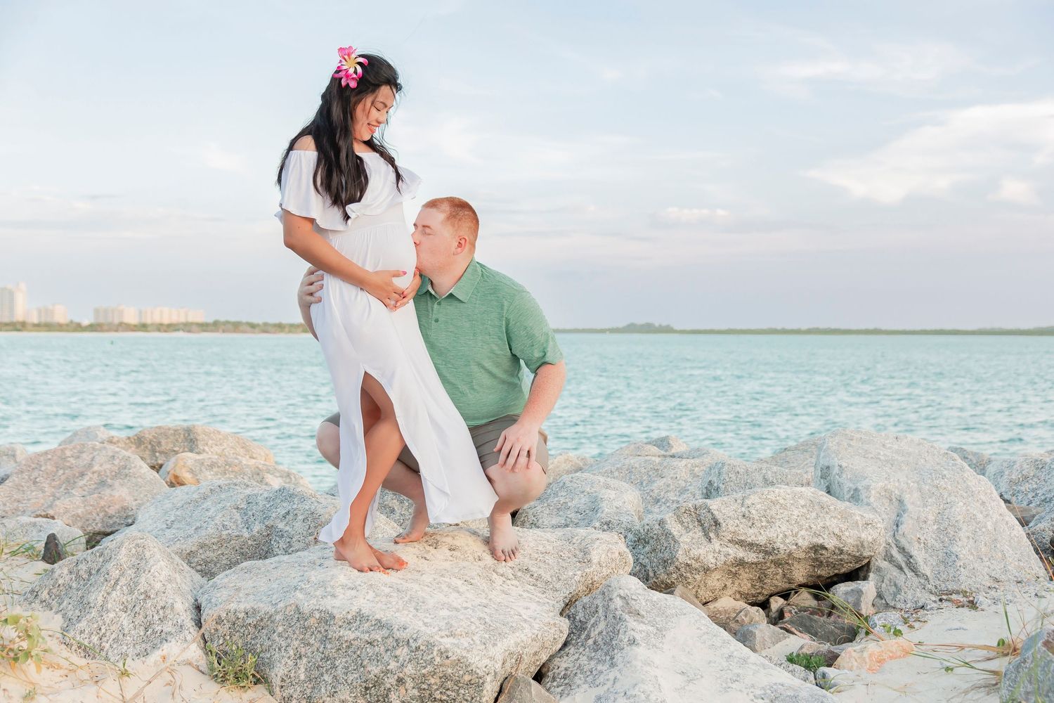 Couple poses on rocky shoreline with turquoise ocean water in background.