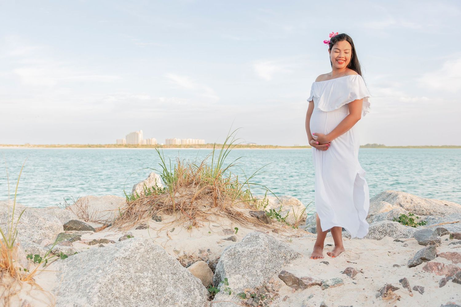 Pregnant woman in white dress poses on rocky shore with turquoise water.