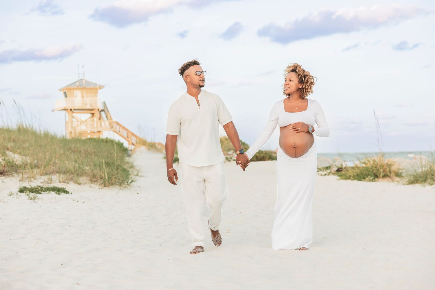 Pregnant couple holding hands walking on a sandy beach near a lifeguard tower at sunset.