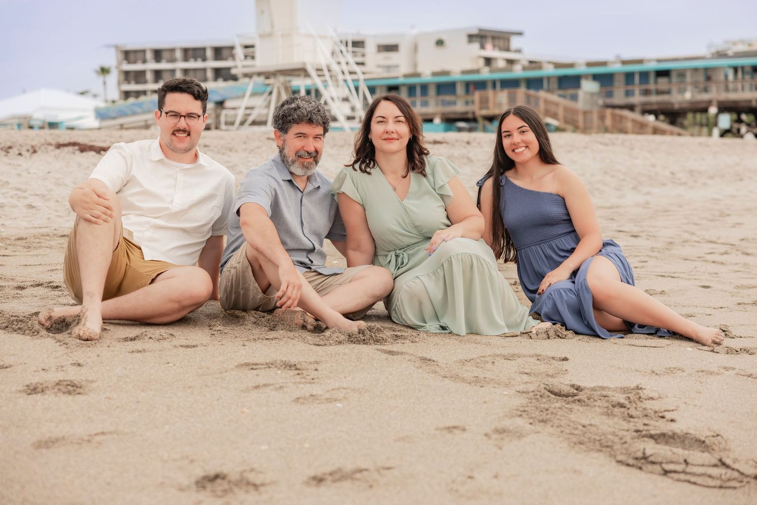 Four adults sitting together on sandy beach with pier and buildings in background.