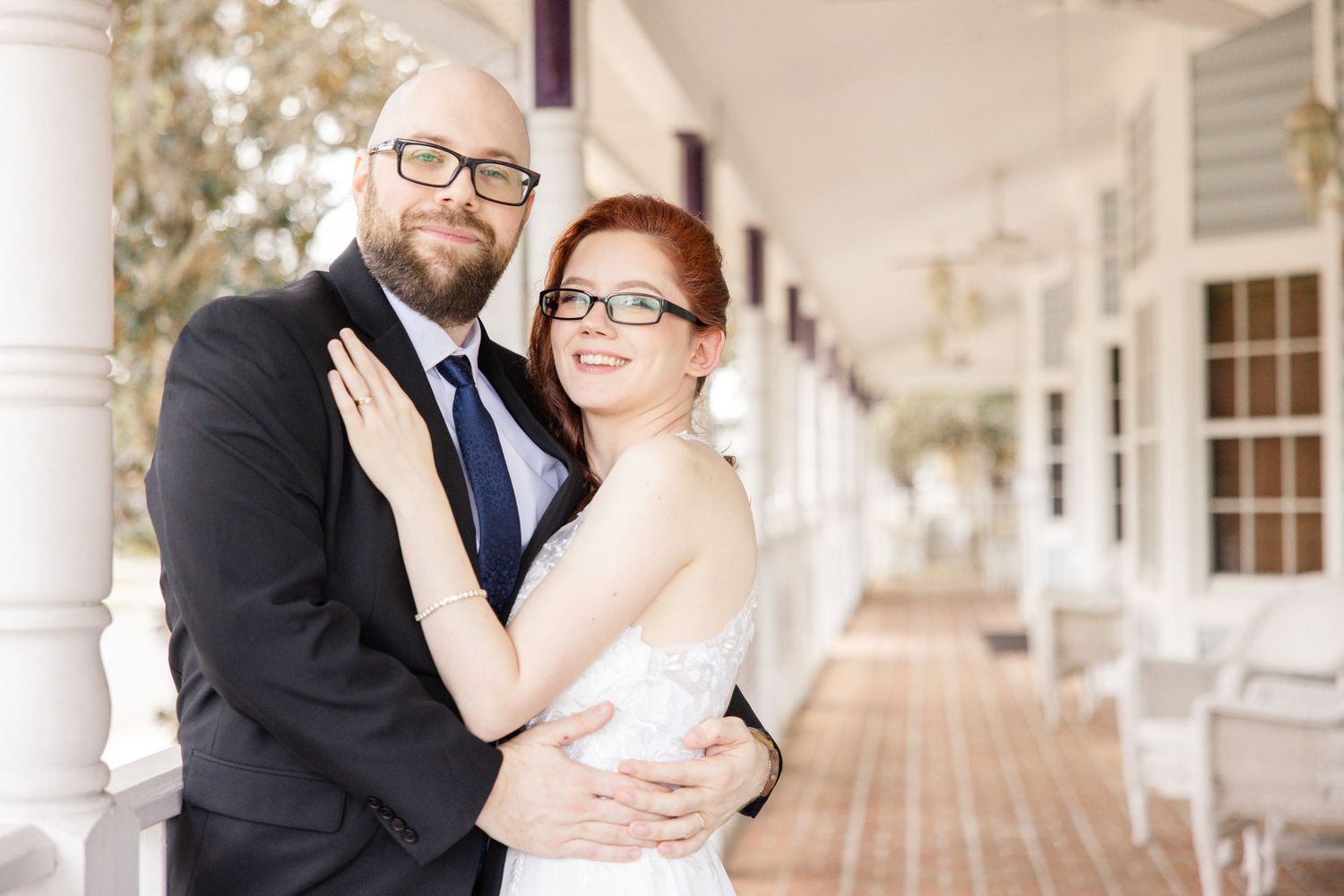 Bearded groom in glasses hugs redhead bride on vintage porch of white building.