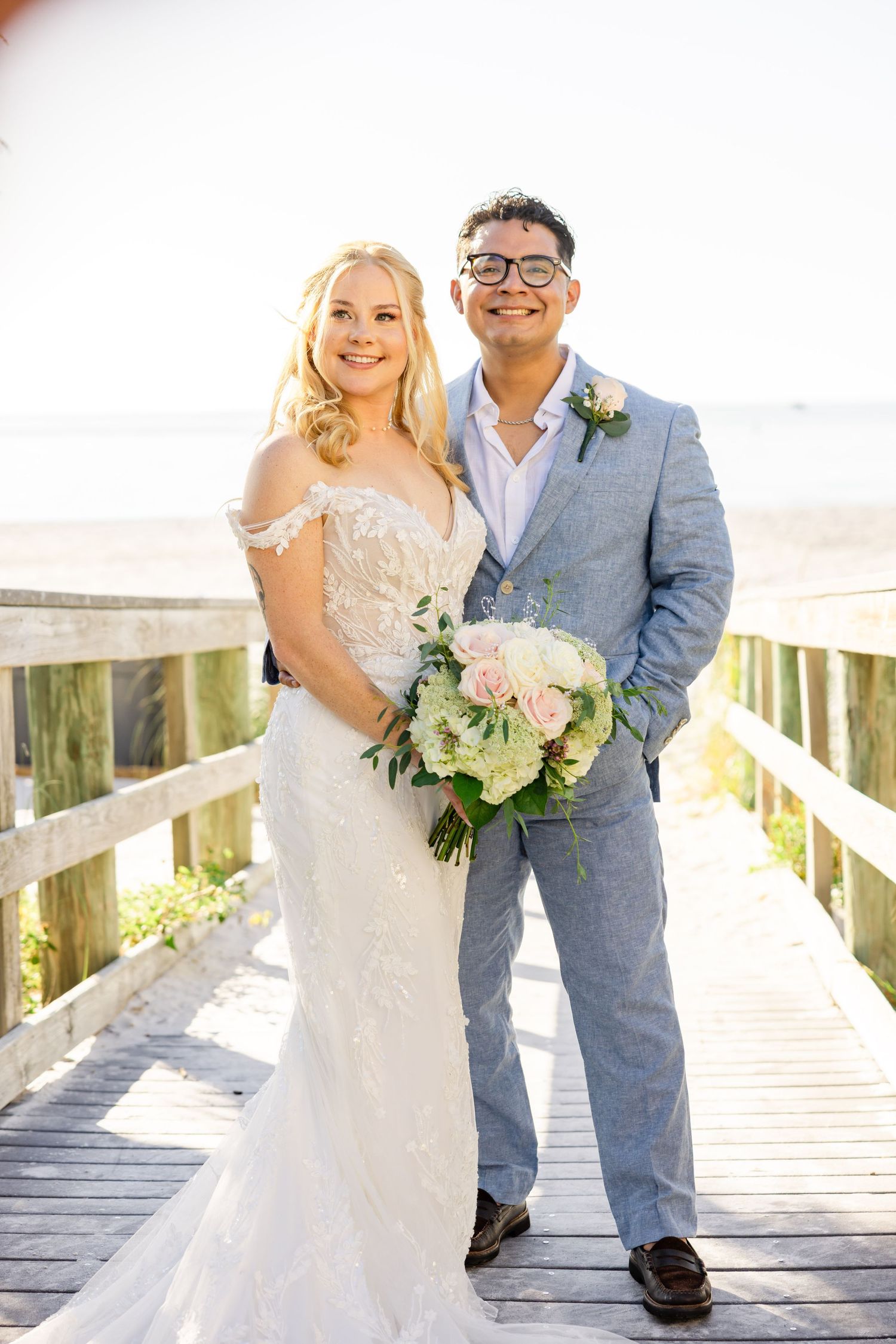 Smiling bride in lace gown and groom in light blue suit stand on beach boardwalk with blush bouquet.