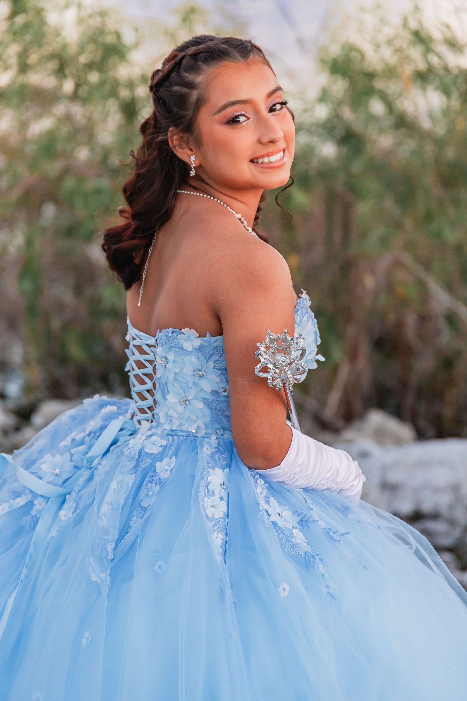 Young woman in light blue quinceañera ball gown with floral appliques smiling outdoors.