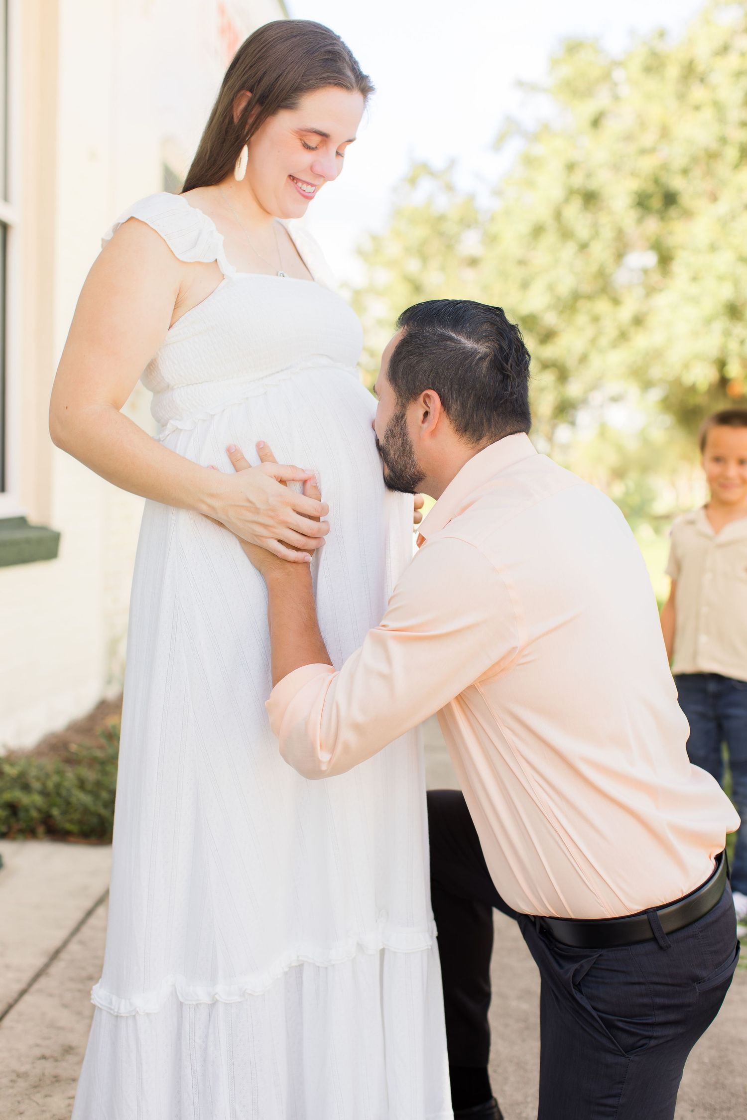 Man kneeling and kissing pregnant woman's baby bump outdoors during maternity photo session.