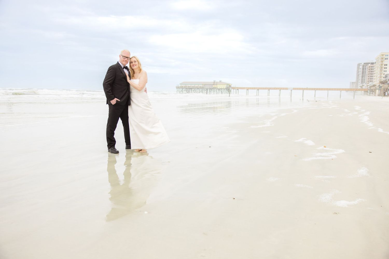 Bride and groom stand barefoot on wet sand near pier during beach wedding portrait.