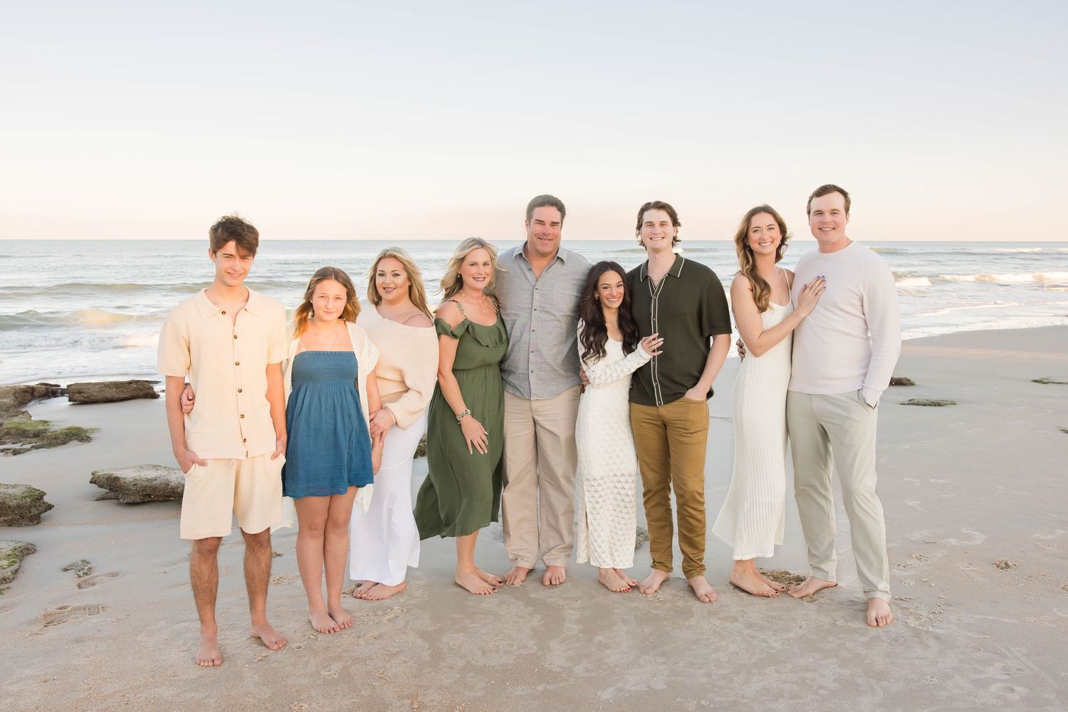 Large family group of eight posing together smiling on a sandy beach at sunset.