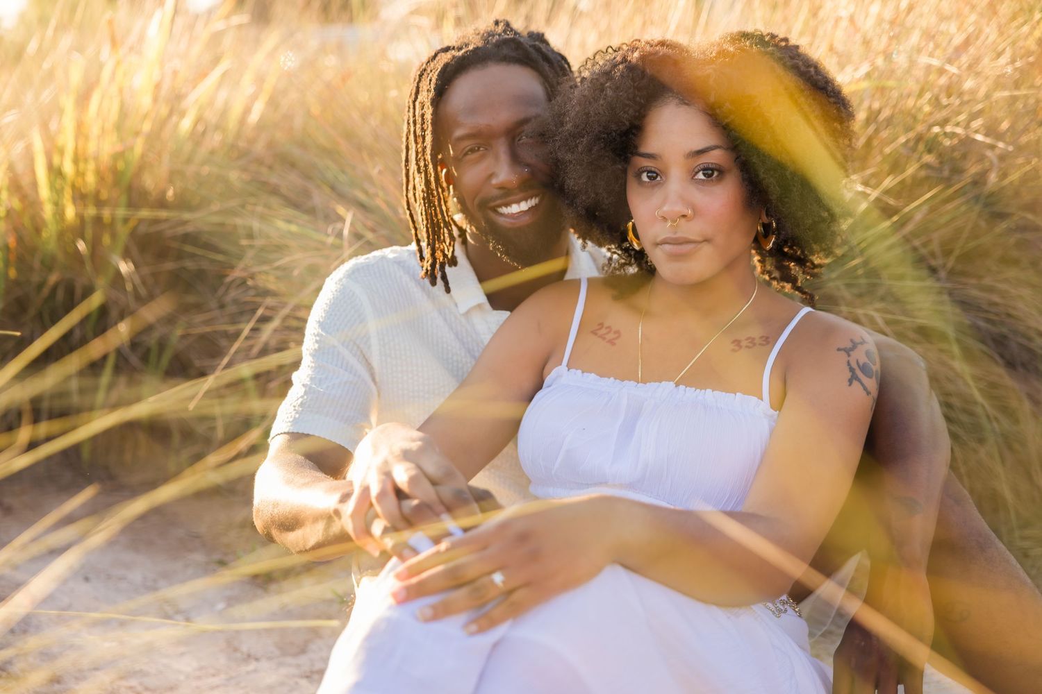 Expectant couple sitting in golden beach grass, man with dreadlocks embracing pregnant woman in white dress.