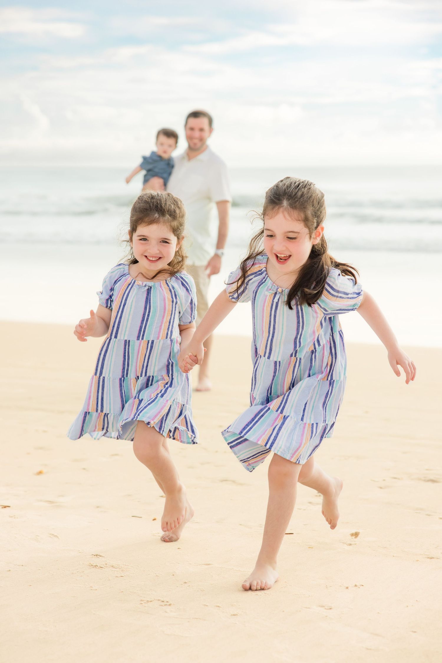 Two girls in striped dresses running on beach with father and baby behind them.