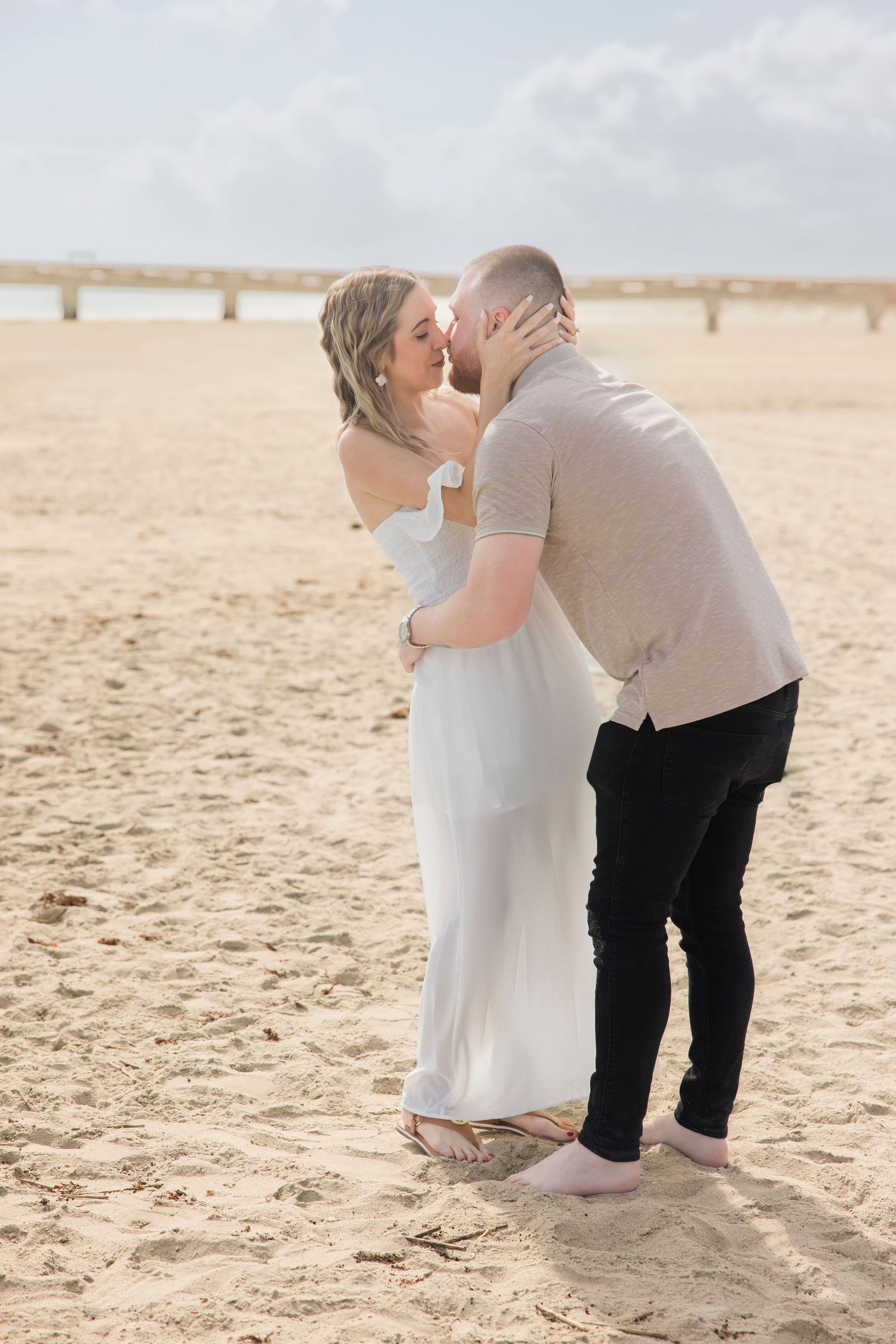 Couple sharing a romantic kiss on a sandy beach, woman in white dress, man in casual attire, pier in background.