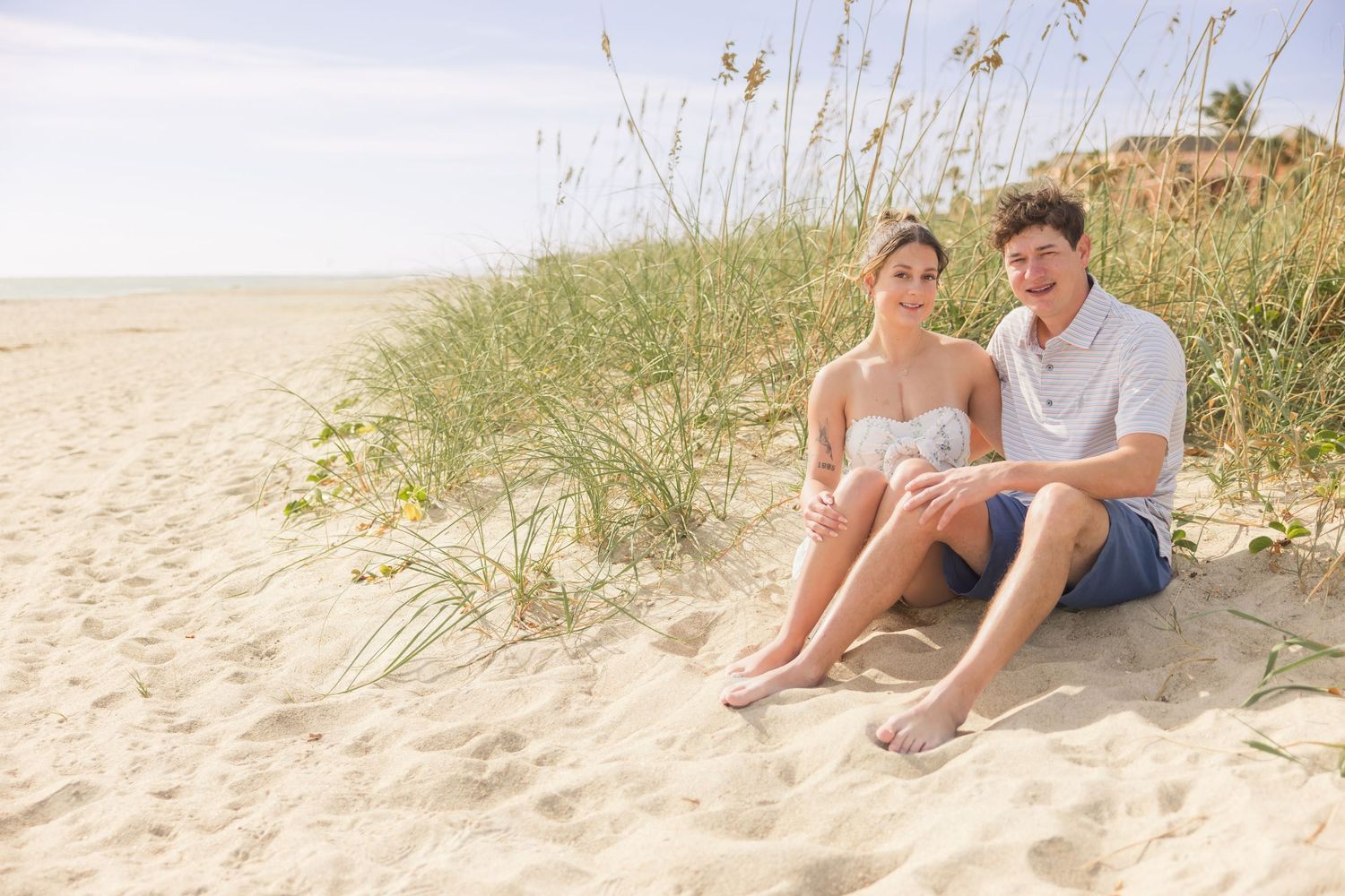 Young couple sitting together on a sandy beach dune surrounded by tall grasses on a sunny day.