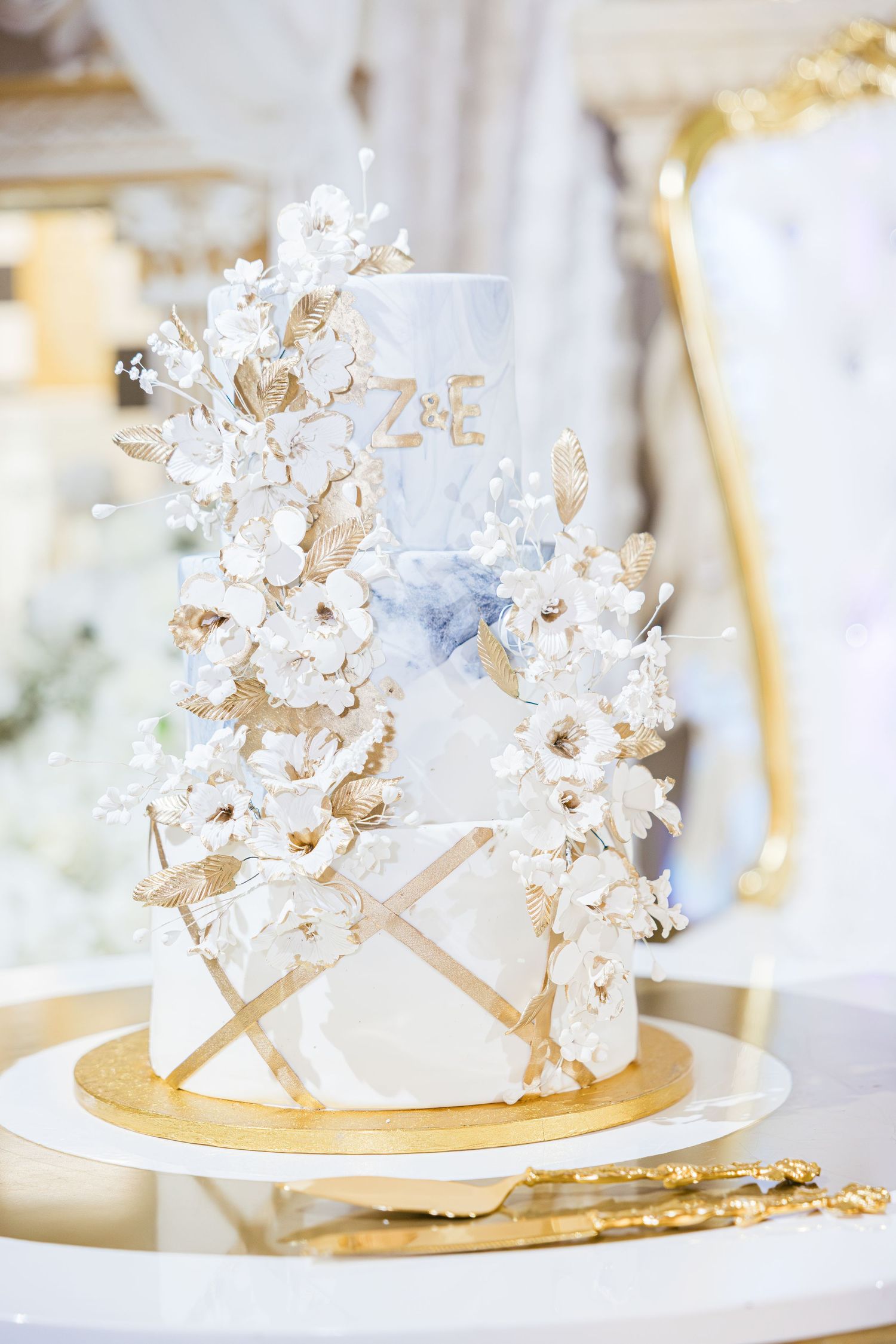 Elegant two-tier white and gold wedding cake adorned with white sugar flowers and gold leaves, monogrammed with Z&E.