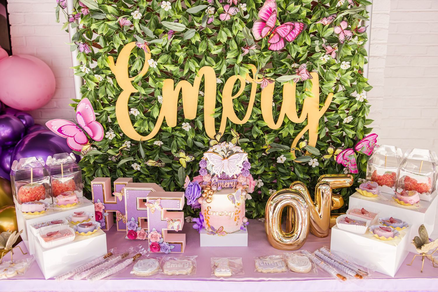 Butterfly-themed birthday dessert table with gold Emery sign and floral backdrop.