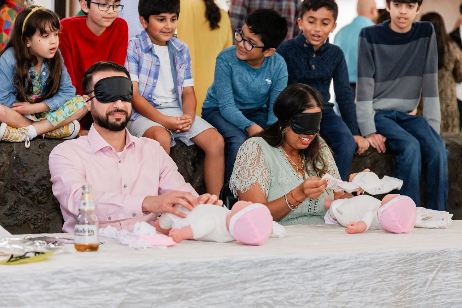 Blindfolded couple diapering baby dolls during a fun baby shower game with kids watching.