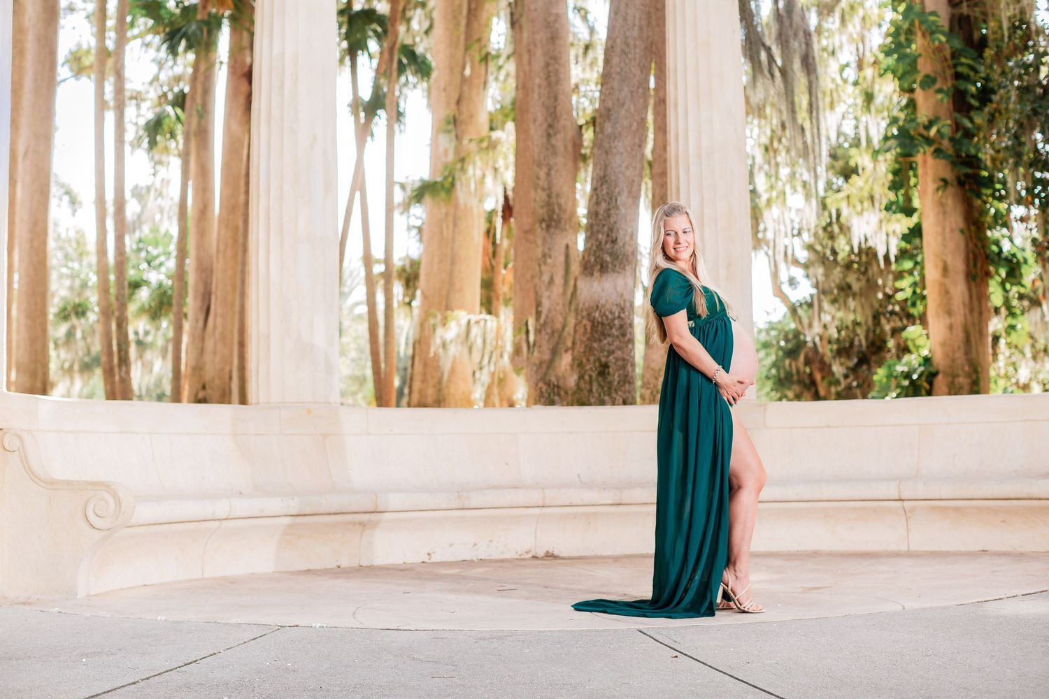 Pregnant woman in emerald gown poses near white columns among tall trees.