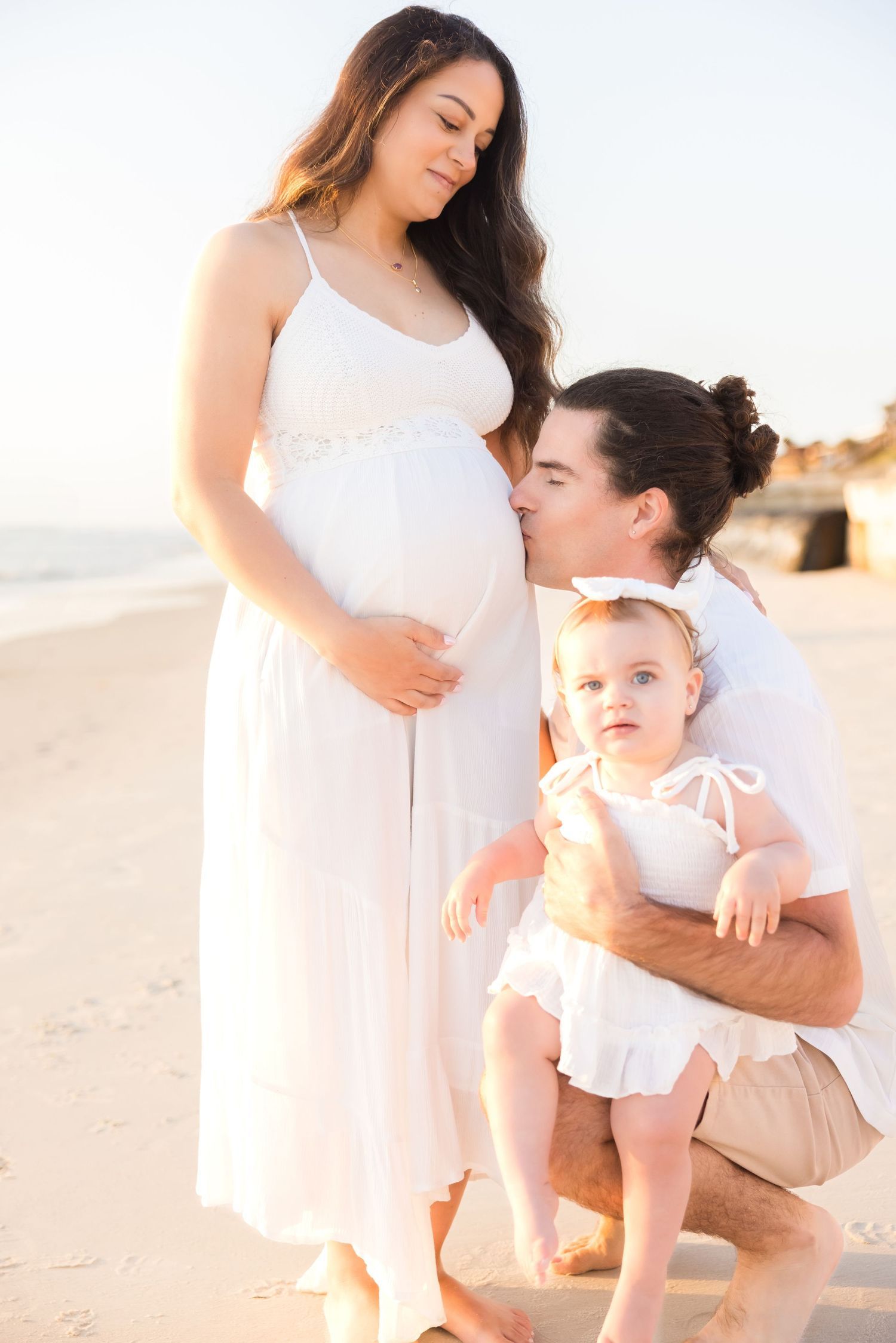 Man kissing pregnant womans belly while holding toddler on a bright sunny beach.