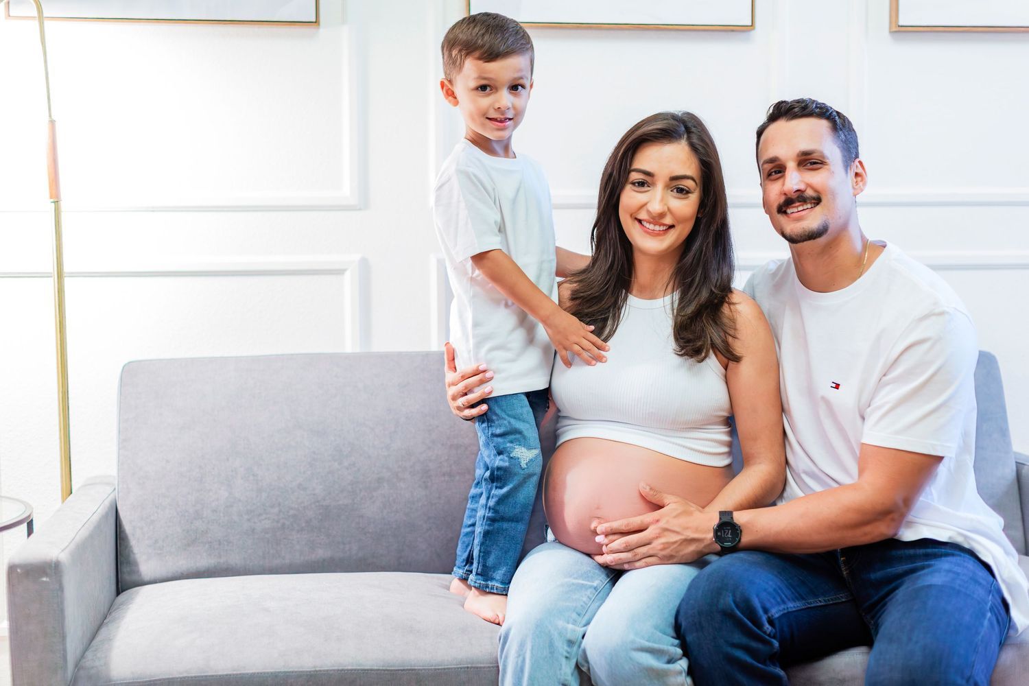 Happy family with pregnant mother, father, and young son posing together on a gray sofa.