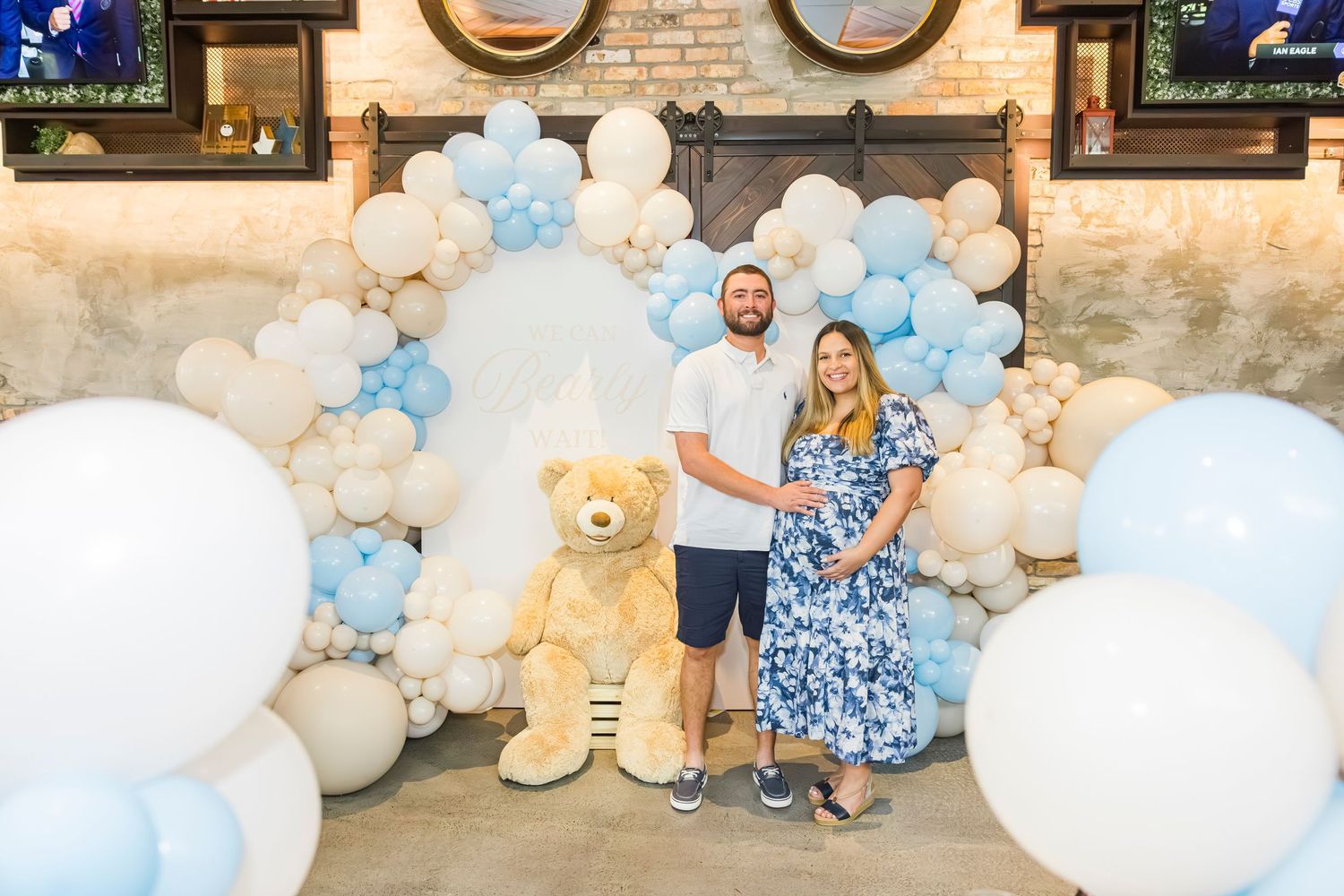 Expecting couple poses with giant teddy bear at blue and white balloon arch baby shower backdrop.