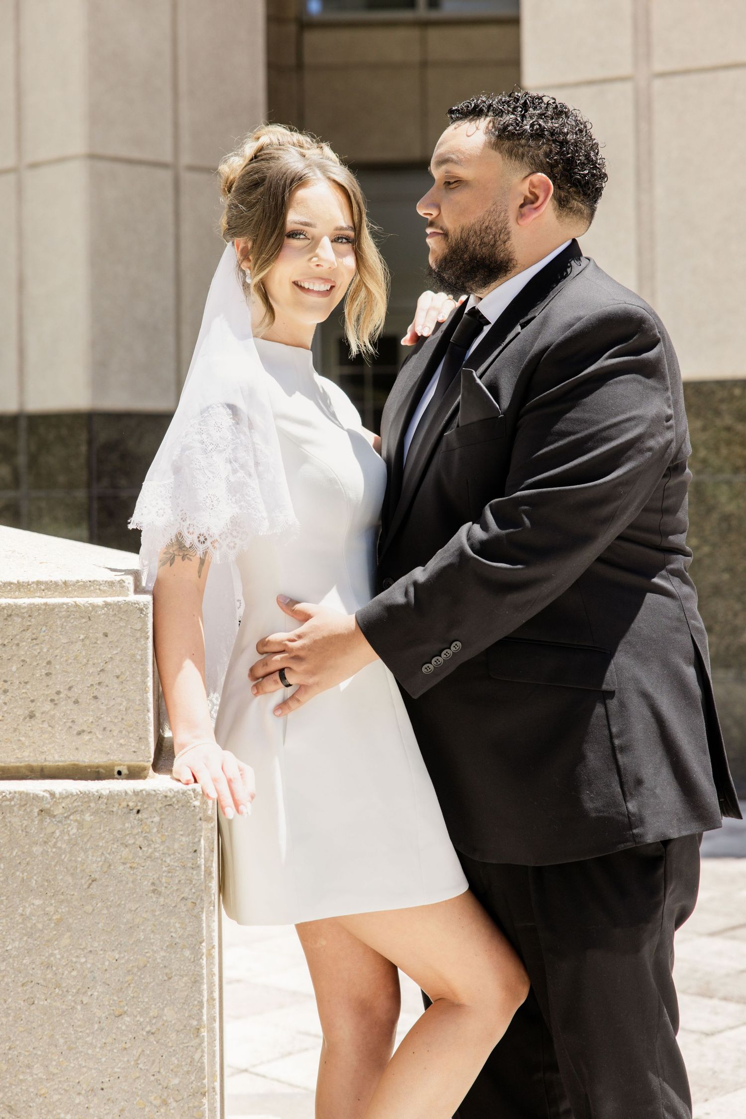 Bride in short white dress and groom in black suit embrace outside building.