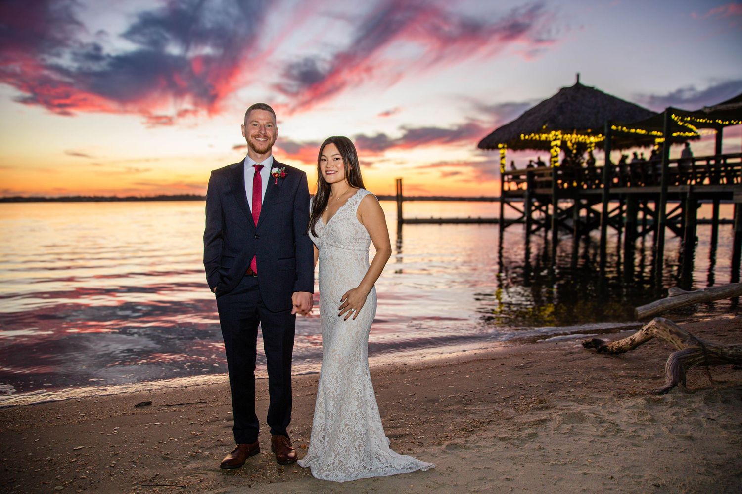 Wedding couple holding hands on beach at dramatic purple and orange sunset with tiki pier backdrop.