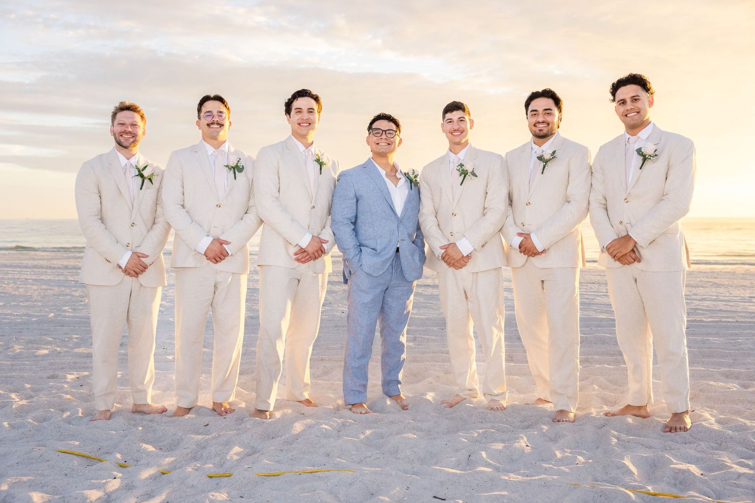 Groom in light blue suit stands with six groomsmen in tan linen suits on sandy beach at sunset.