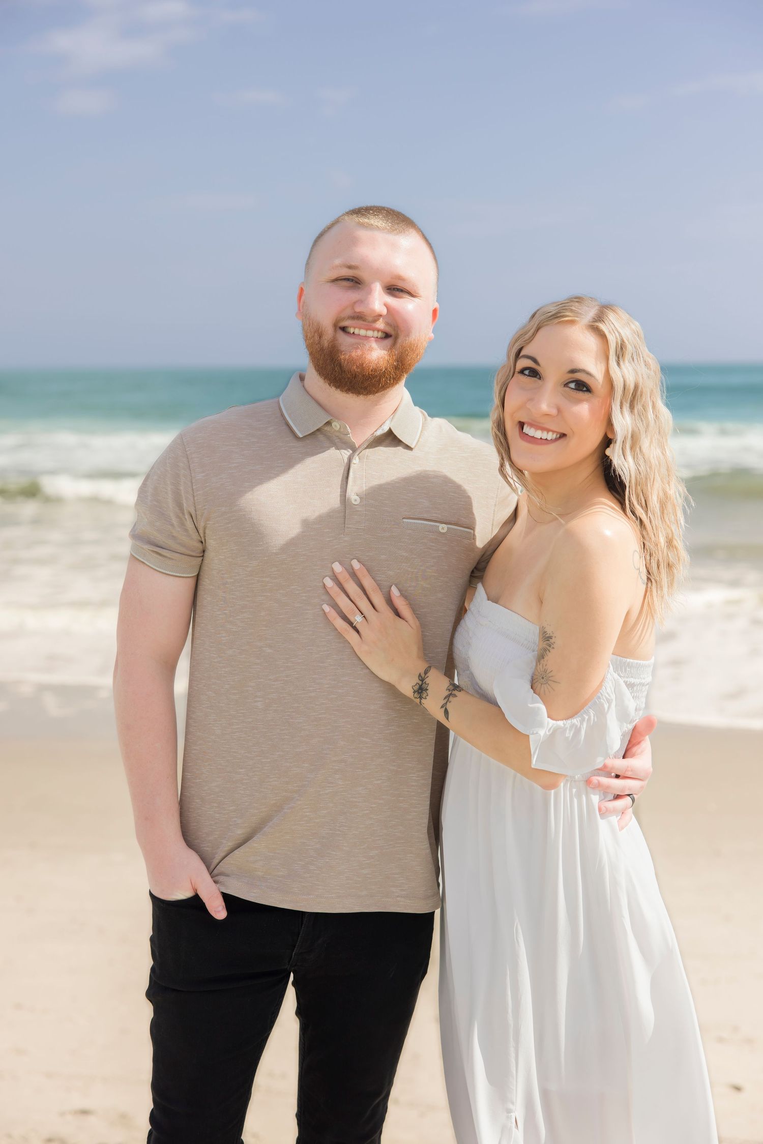 Happy couple posing together on a sunny beach, woman in white dress showing engagement ring, man in beige polo shirt.