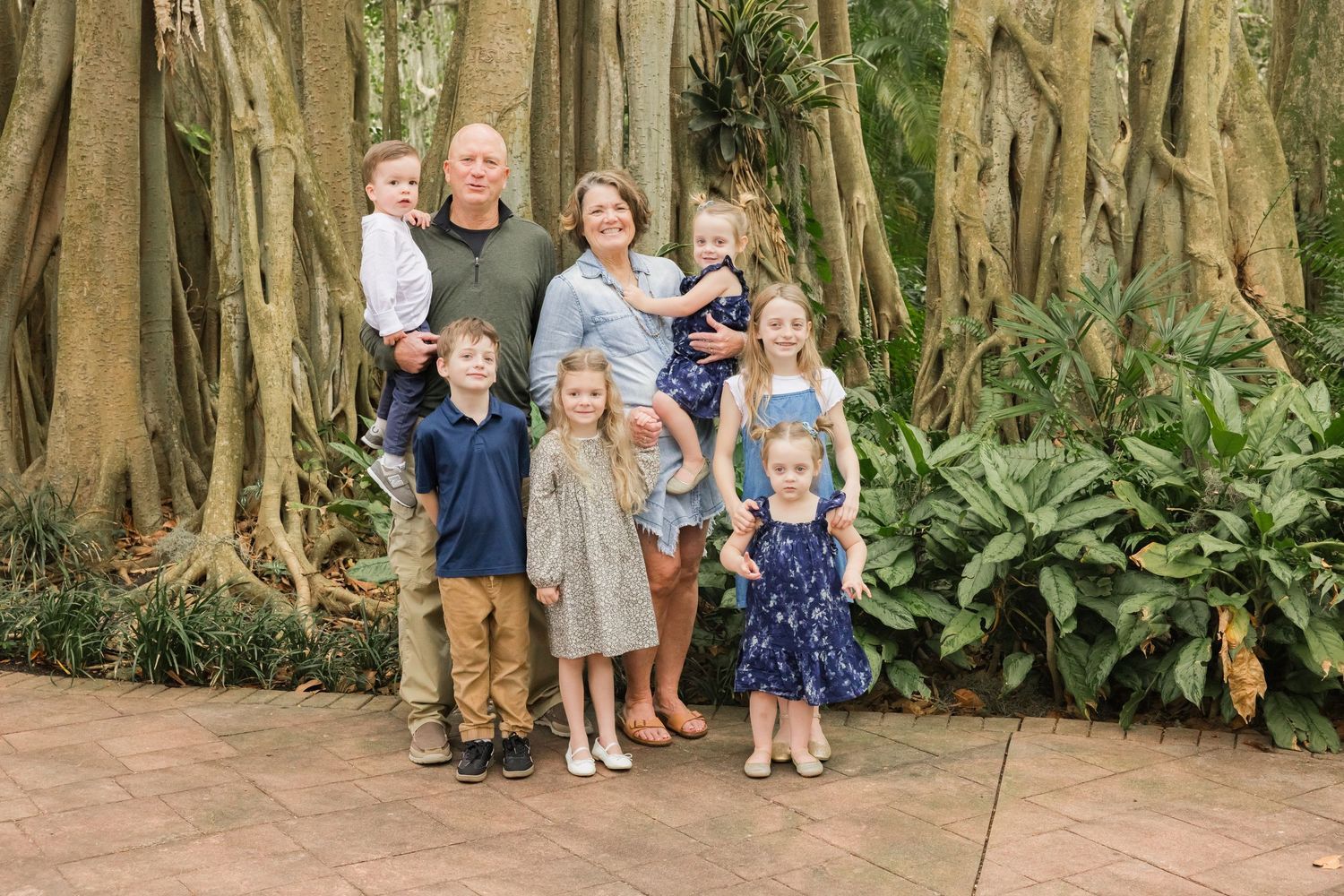 Grandparents posing with six grandchildren in front of a large banyan tree in a tropical garden.