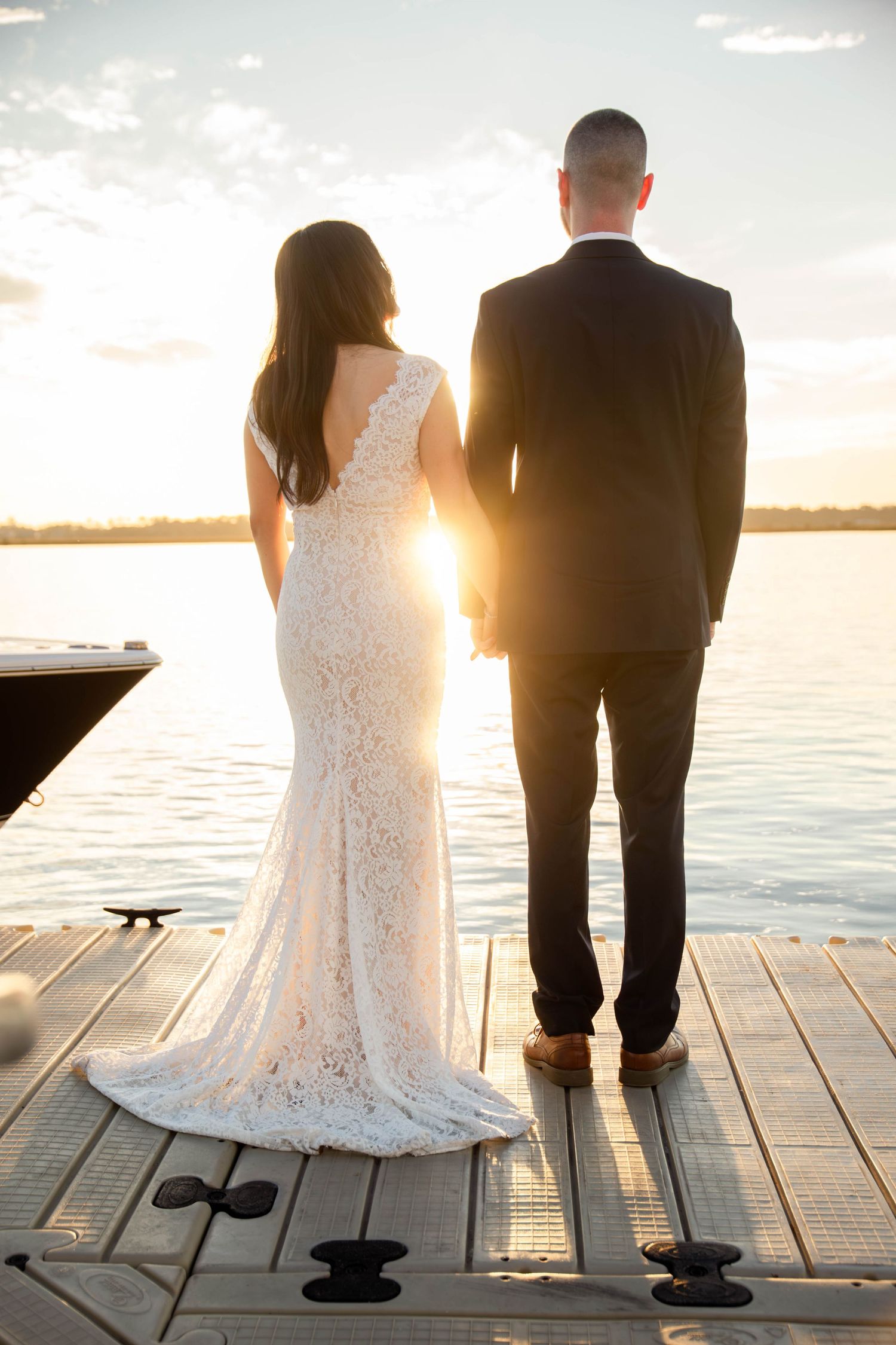 Bride and groom holding hands on a dock at sunset, overlooking a calm lake, viewed from behind.