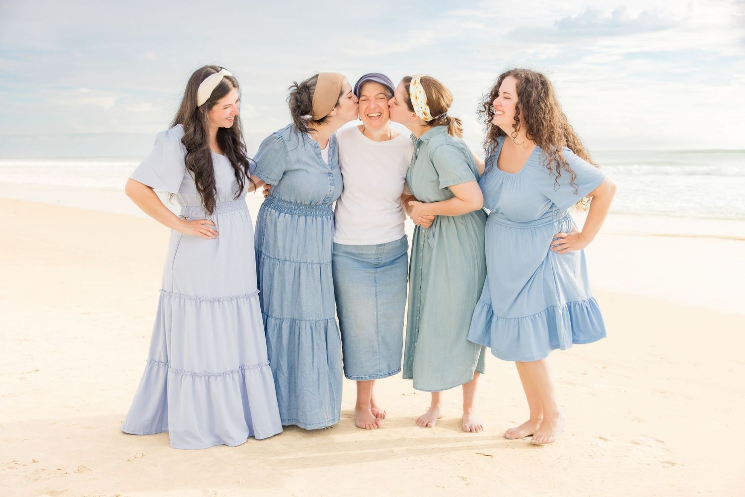 Five women in blue dresses laughing together on a sunny beach.