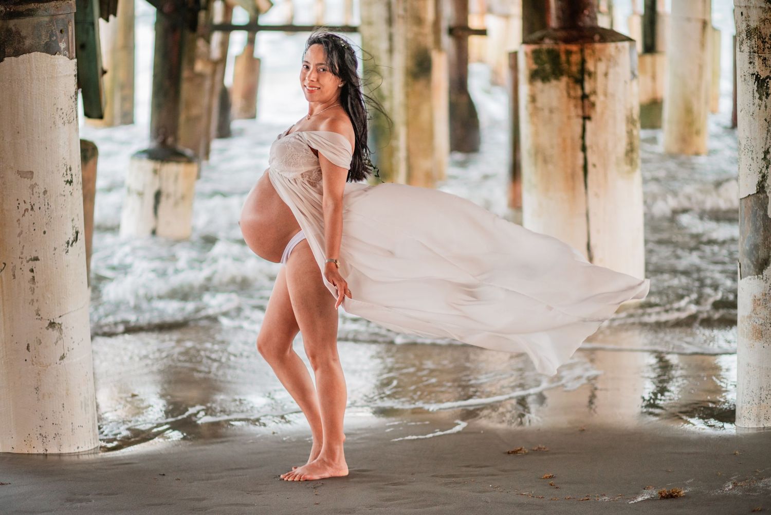 Pregnant woman in flowing white skirt posing barefoot under pier with ocean waves at beach.