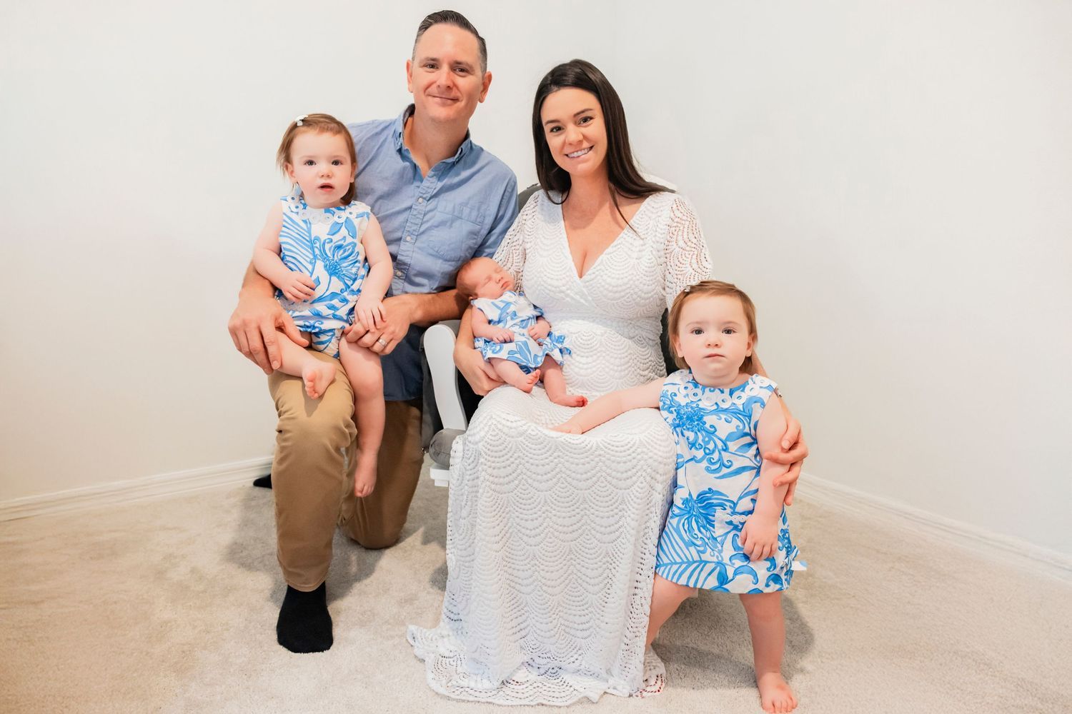 Happy family of five posing together, parents with two toddlers and a newborn baby in a bright indoor setting.
