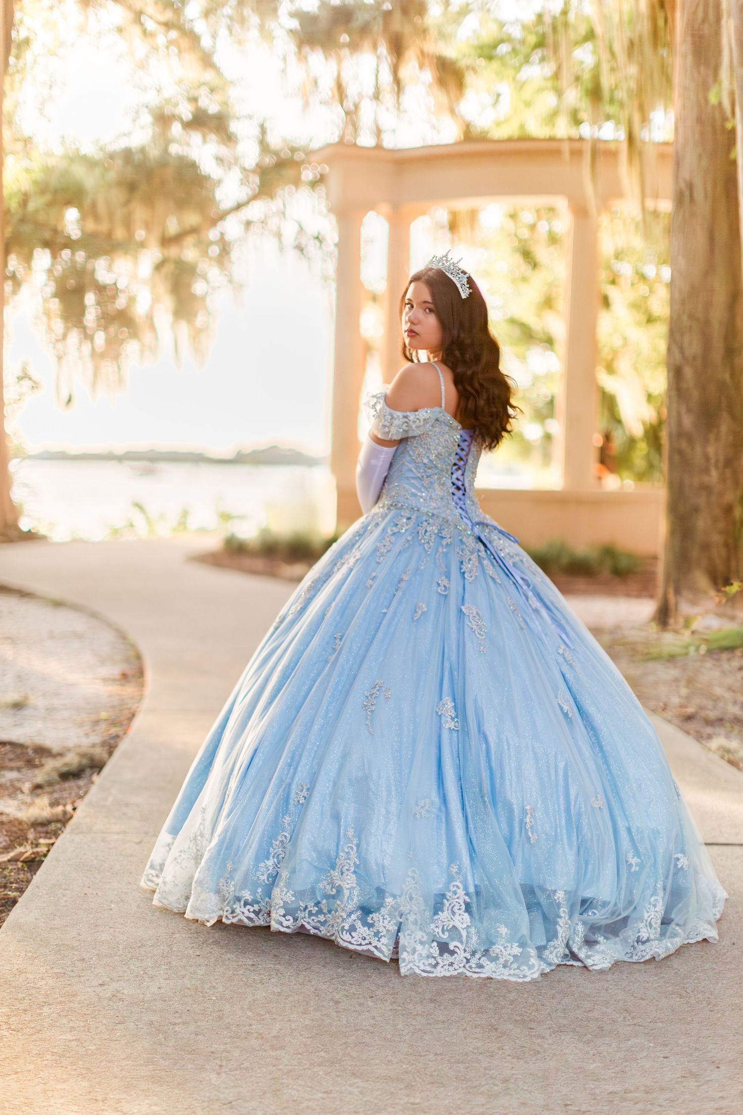 Young woman in a light blue quinceañera ball gown with lace details poses outdoors near a gazebo.