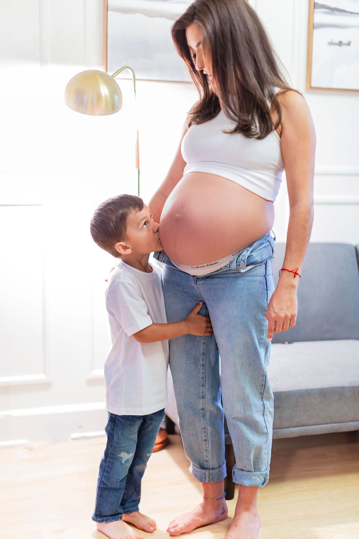 Young boy kissing his pregnant mother's baby bump in a bright home setting.