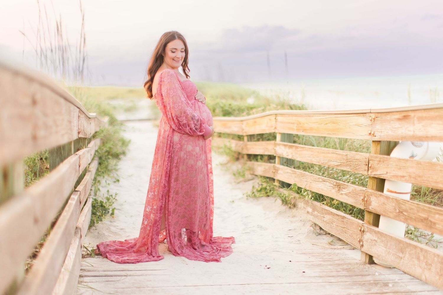 Pregnant woman in flowing pink lace dress standing gracefully on a beach boardwalk at dusk.