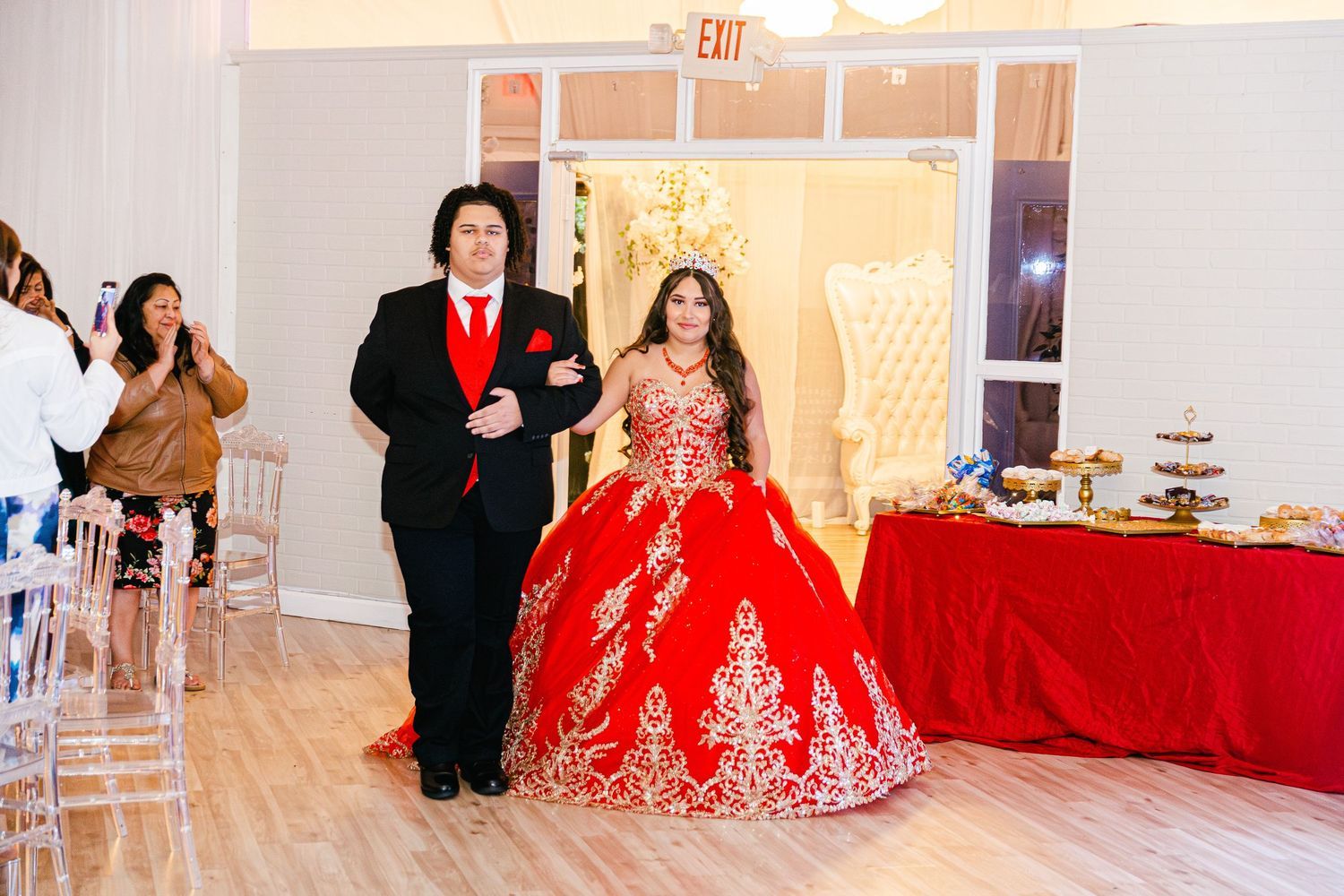 Young woman in red quinceañera ball gown with gold embroidery making grand entrance with escort.