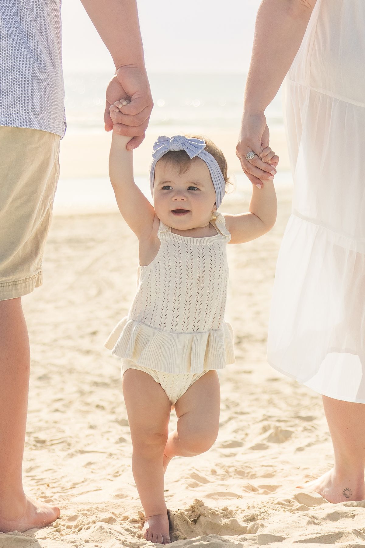 Baby girl in knit dress with bow headband learning to walk holding parents hands on beach.