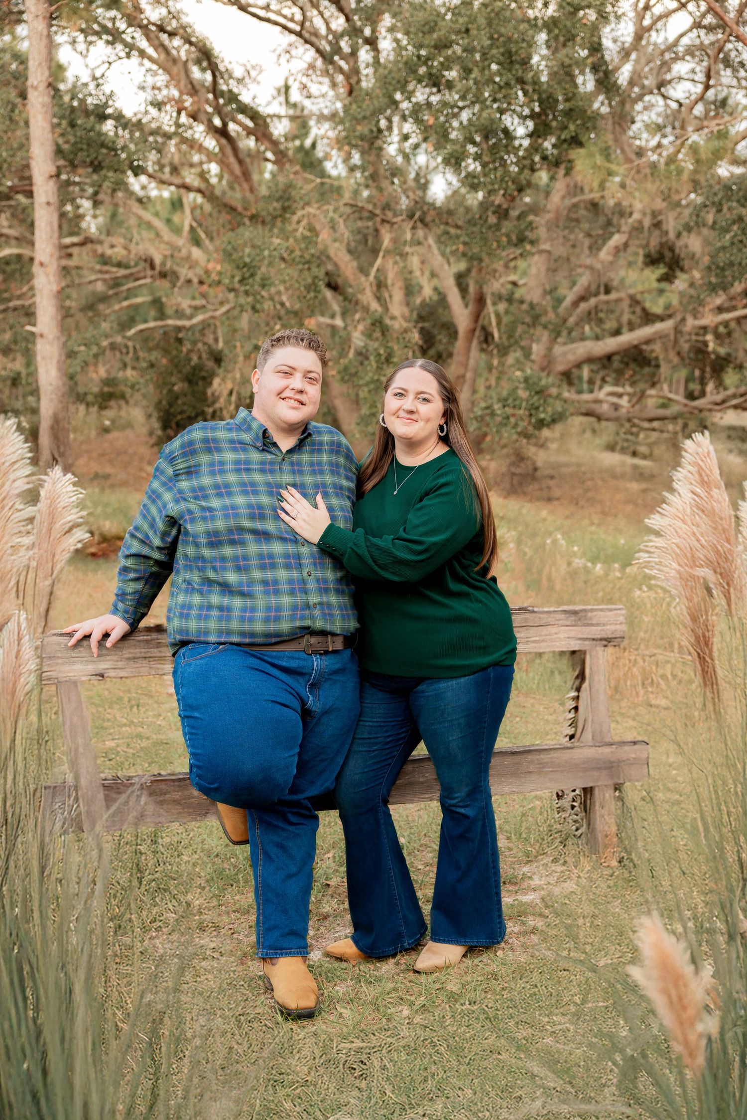 Smiling couple posing against a wooden fence surrounded by pampas grass in a natural setting.