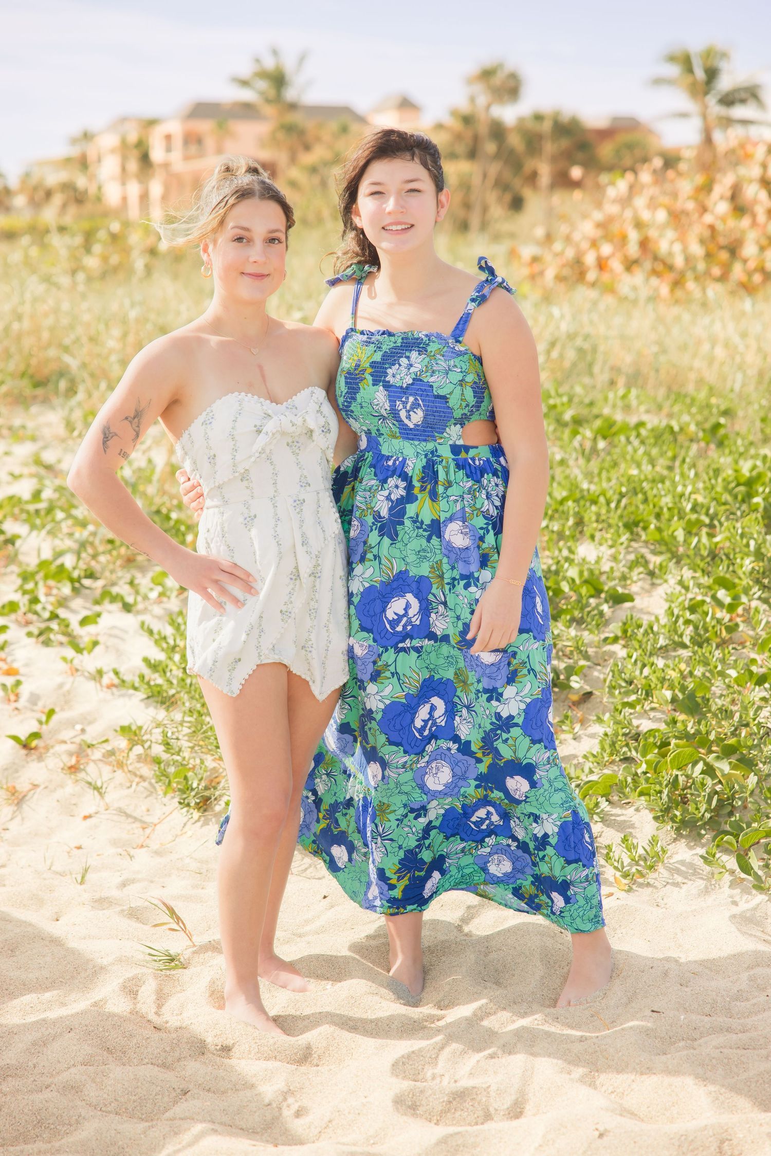 Two young women posing together on a sandy beach, one in a white romper and one in a blue floral dress.