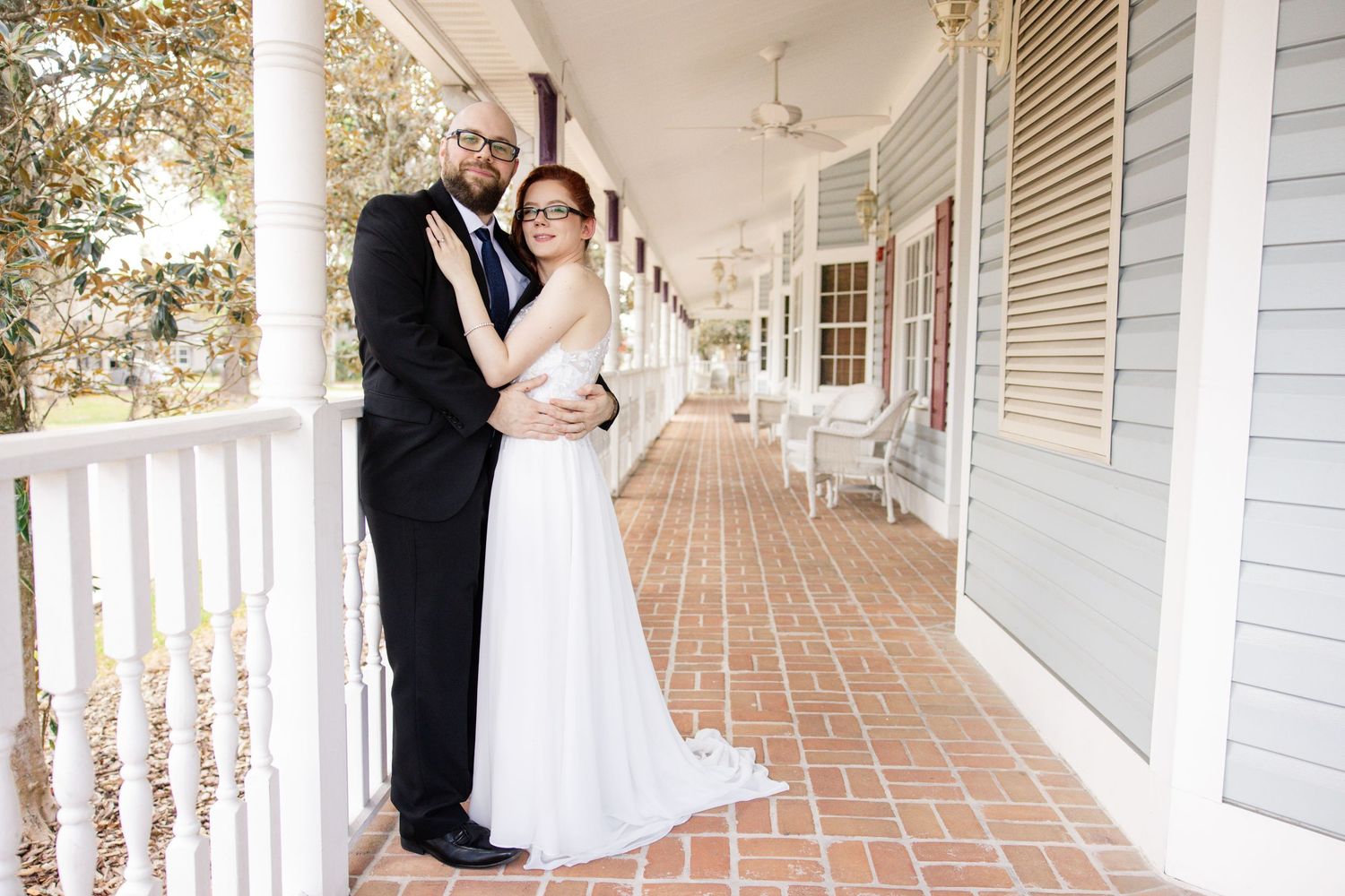 Bride and groom embrace on brick porch of historic white colonial-style building.