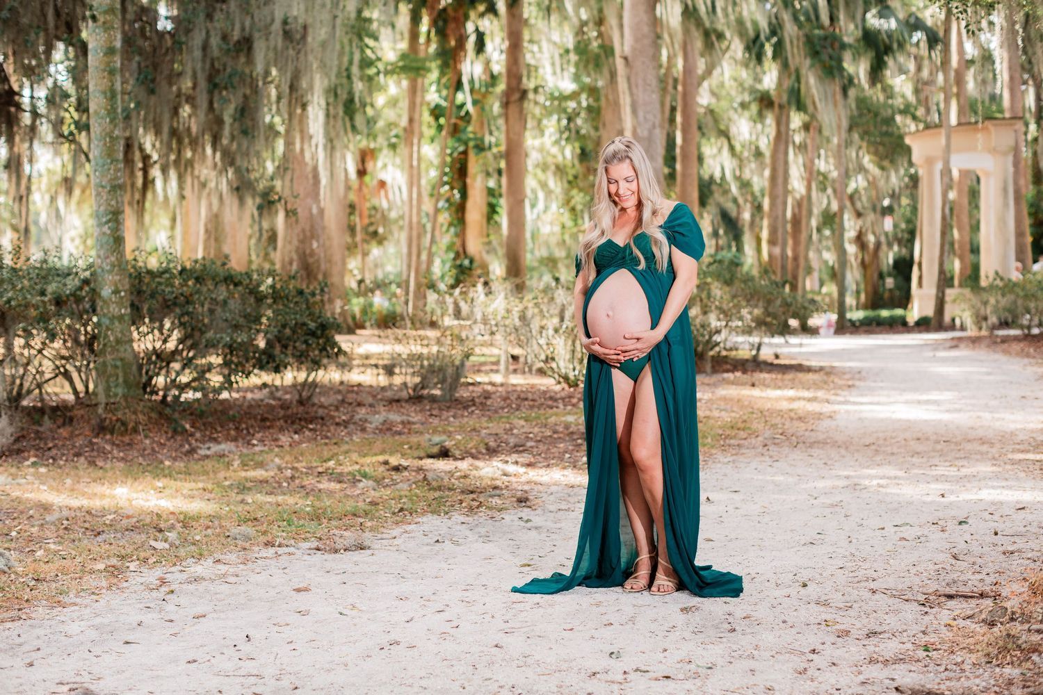 Pregnant woman in emerald green gown poses among tall trees on dirt path.
