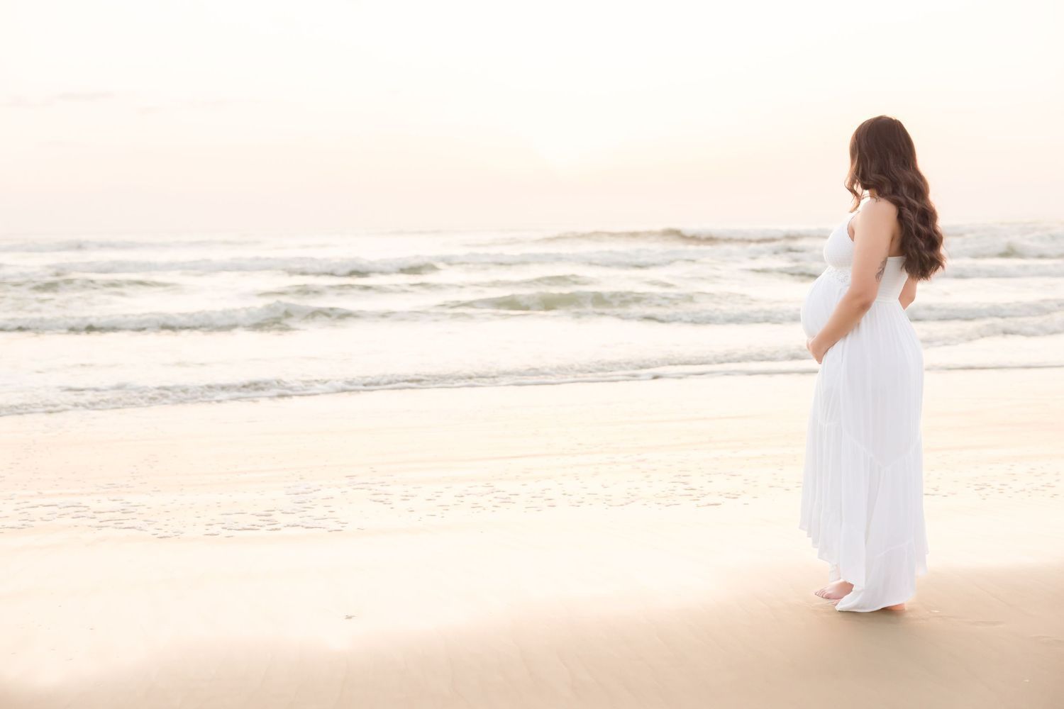 Pregnant woman in white dress standing alone on a serene beach gazing at ocean waves.