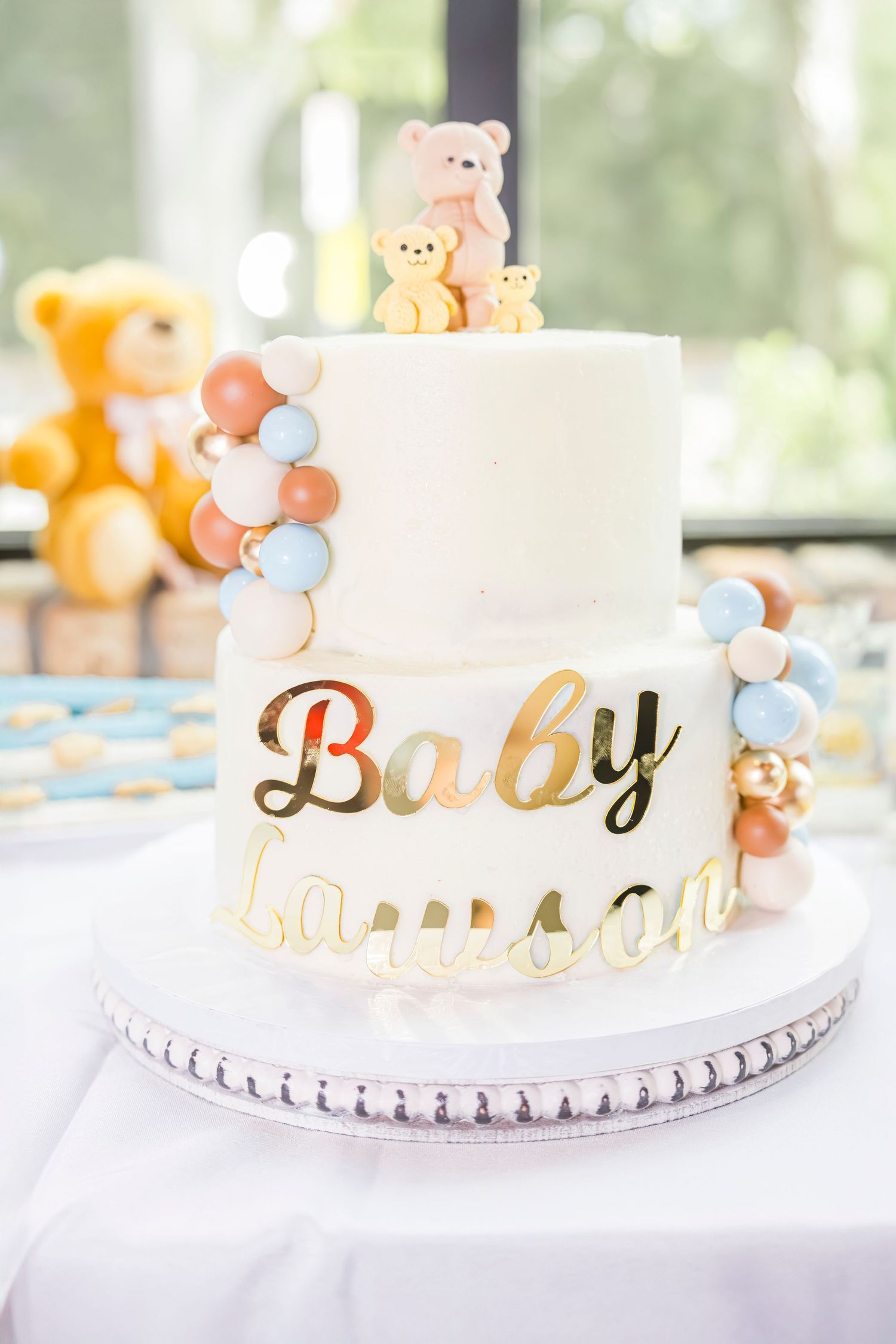 A two-tier white baby shower cake decorated with mini balloons and teddy bears reading Baby Lawson in gold letters.