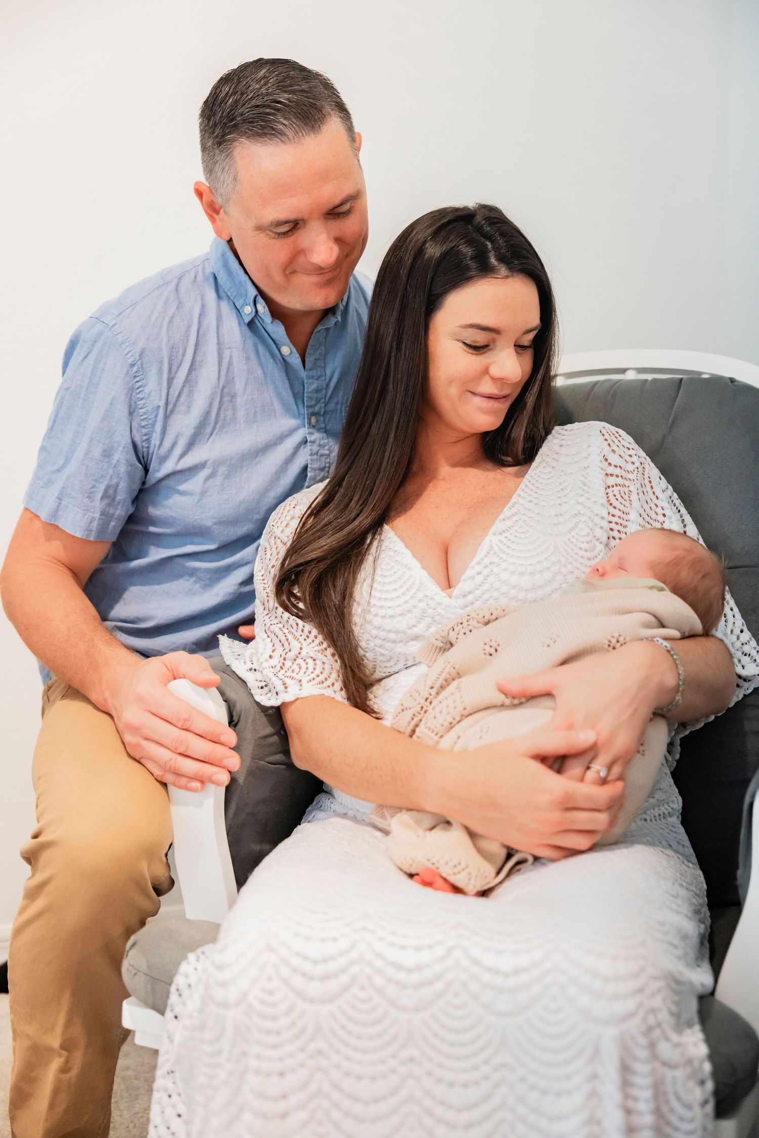 Loving couple admiring their newborn baby, mother in white lace dress seated in a gray chair with father beside her.