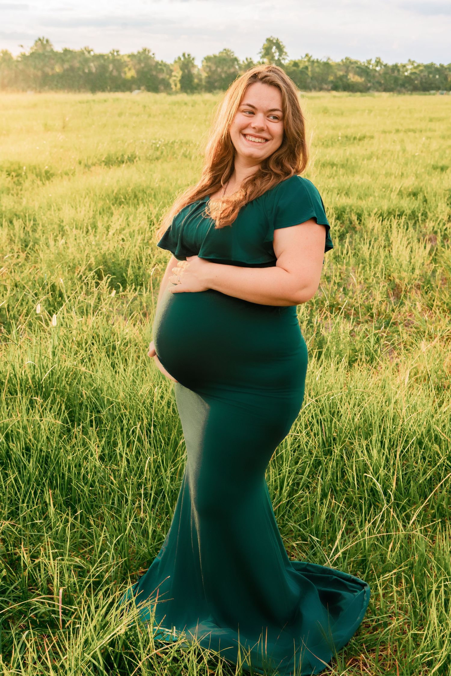 Smiling pregnant woman in green mermaid gown cradling baby bump in lush grass field at golden hour.
