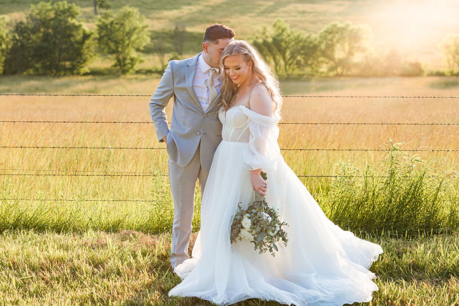 Bride and groom embracing in golden sunset light beside rustic fence in green countryside field.