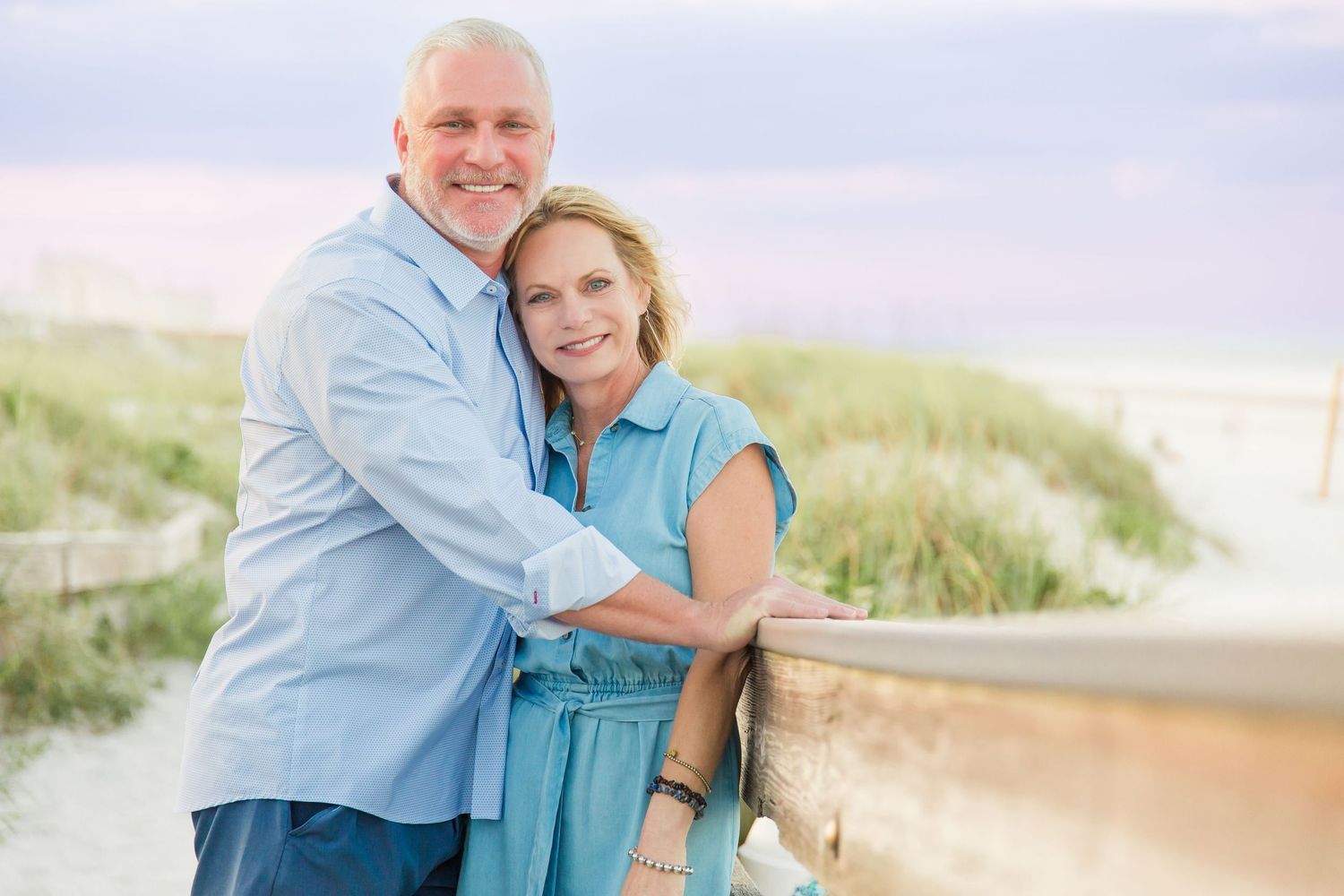 Smiling middle-aged couple embracing near a wooden railing with beach dunes in the background.