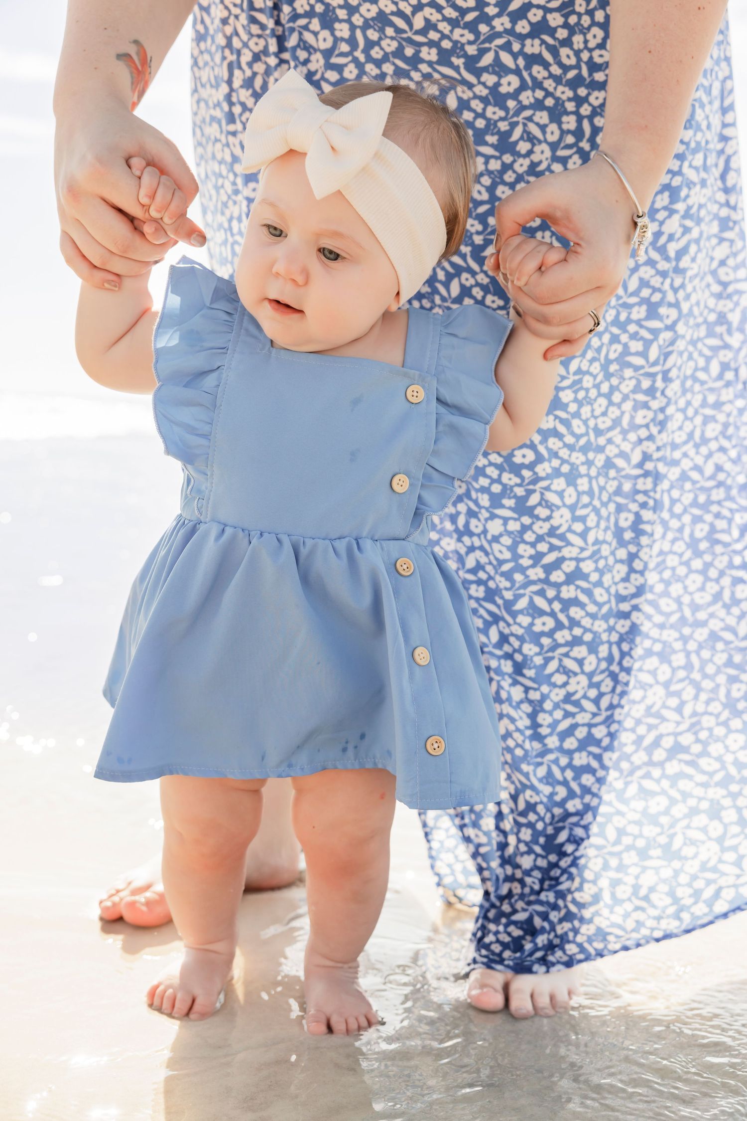 Baby girl in blue dress taking first steps on beach held by mother.