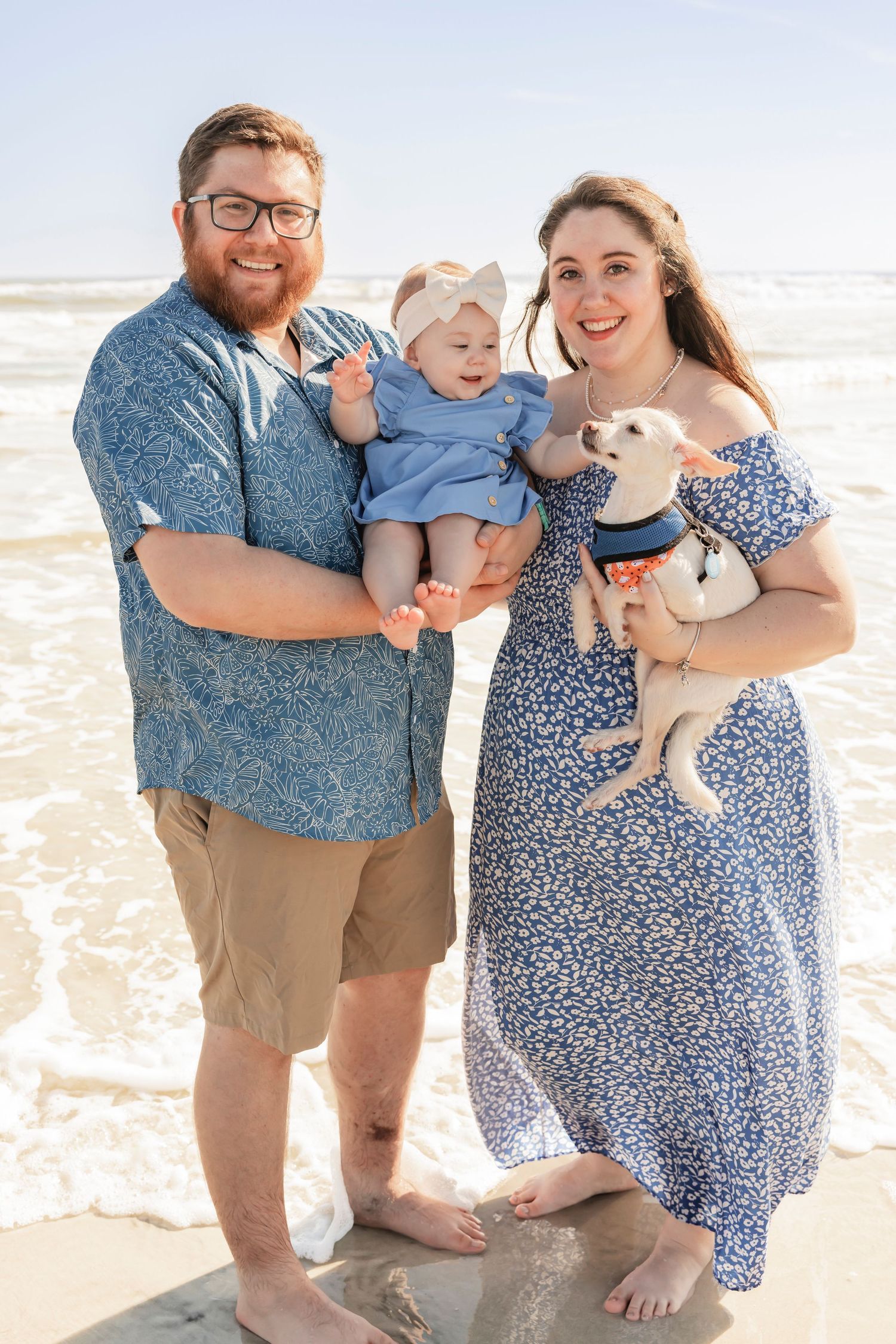 Couple standing on beach holding baby girl and small white dog.