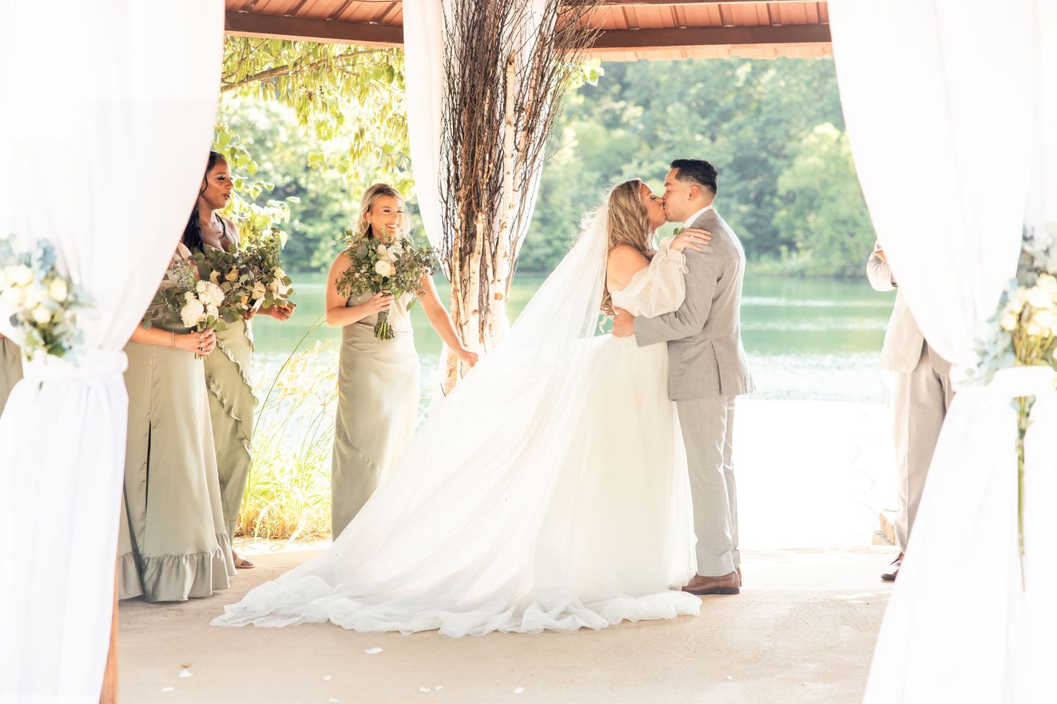Bride and groom share first kiss at outdoor waterfront wedding ceremony with bridesmaids.